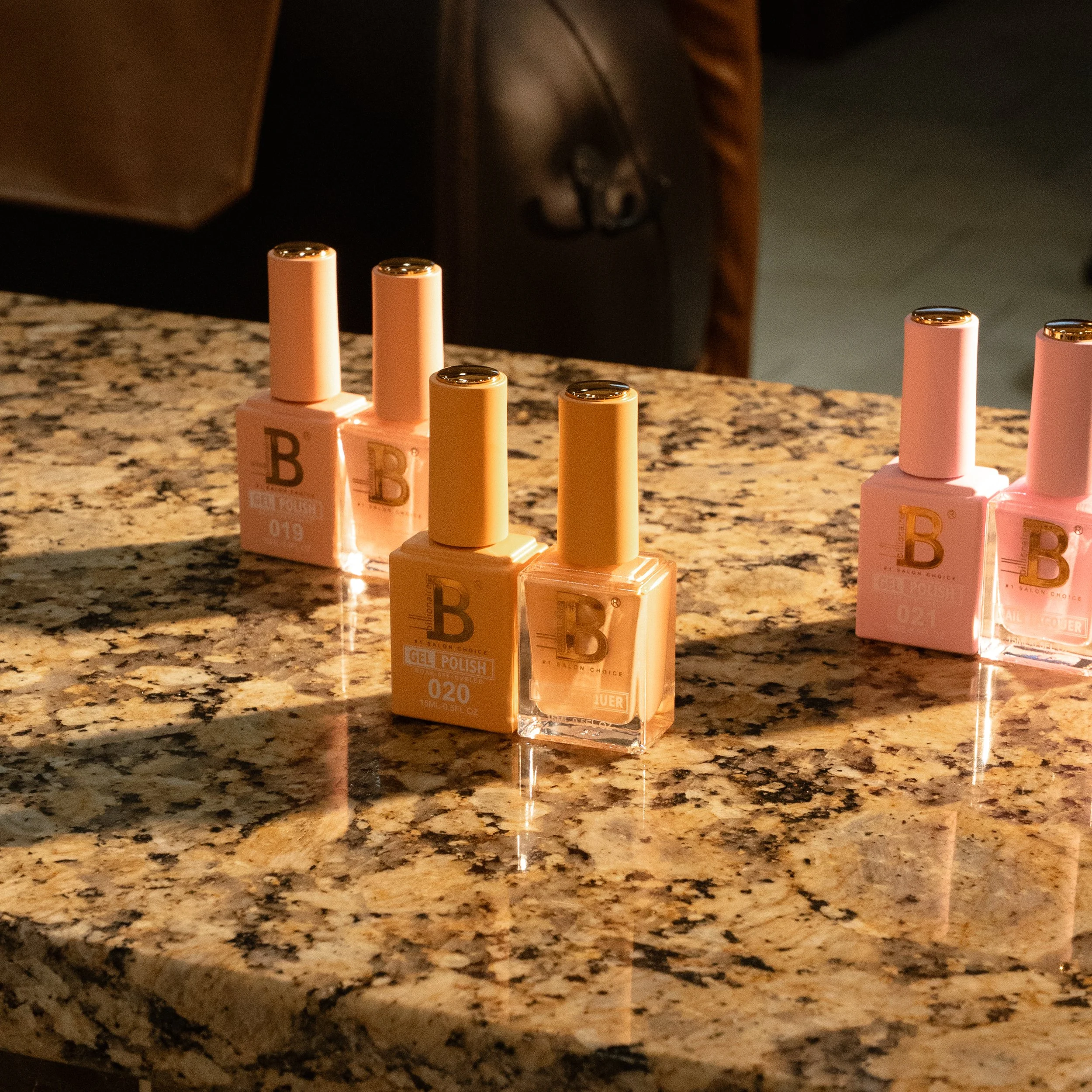 Four bottles of gel nail polish arranged on a speckled granite countertop, with shadows cast by warm lighting. The bottles are in light pink and orange shades.