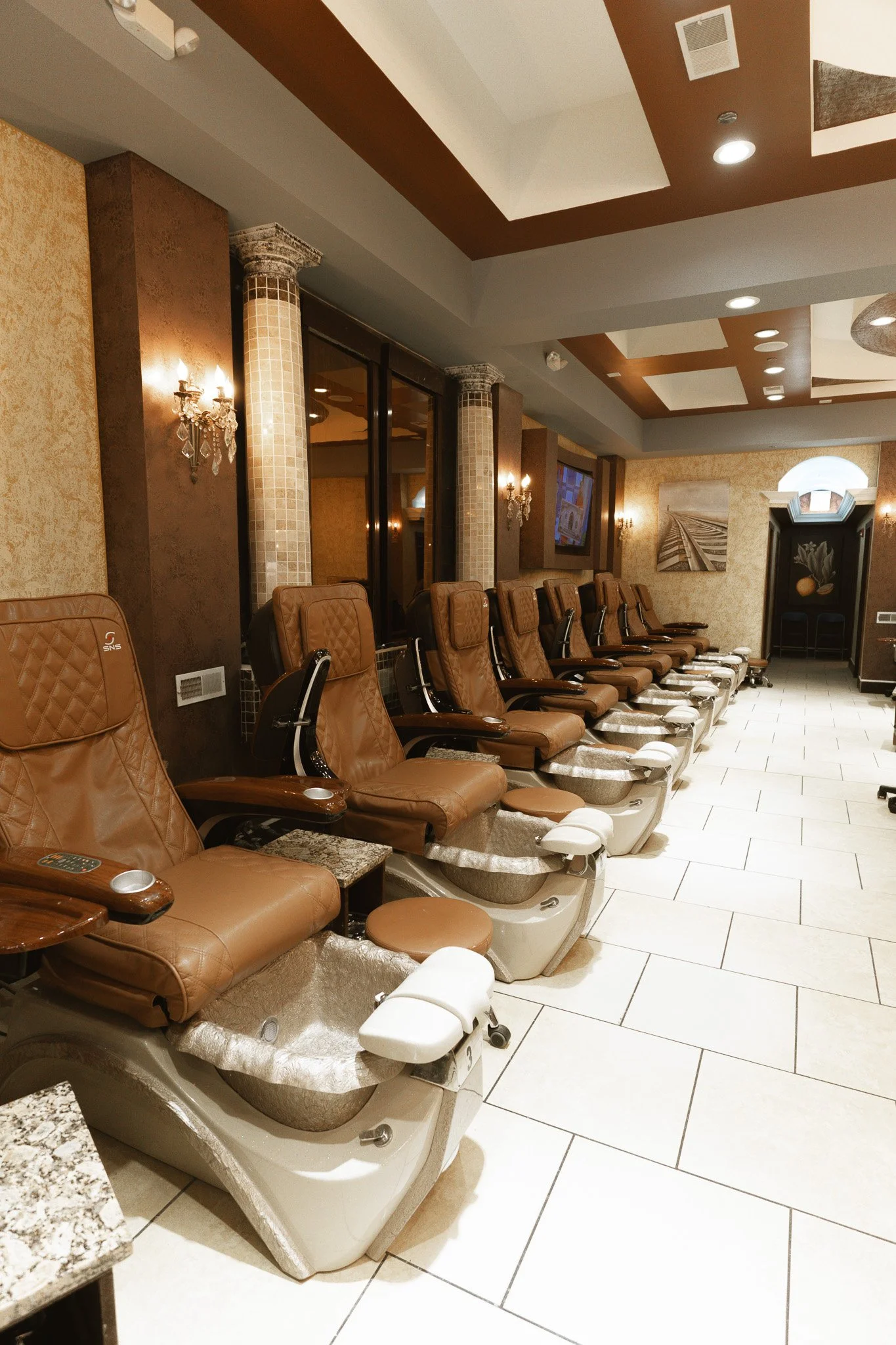 Line of brown massage chairs in a salon, with foot baths underneath, in a warmly lit room with beige tiles and wall art.