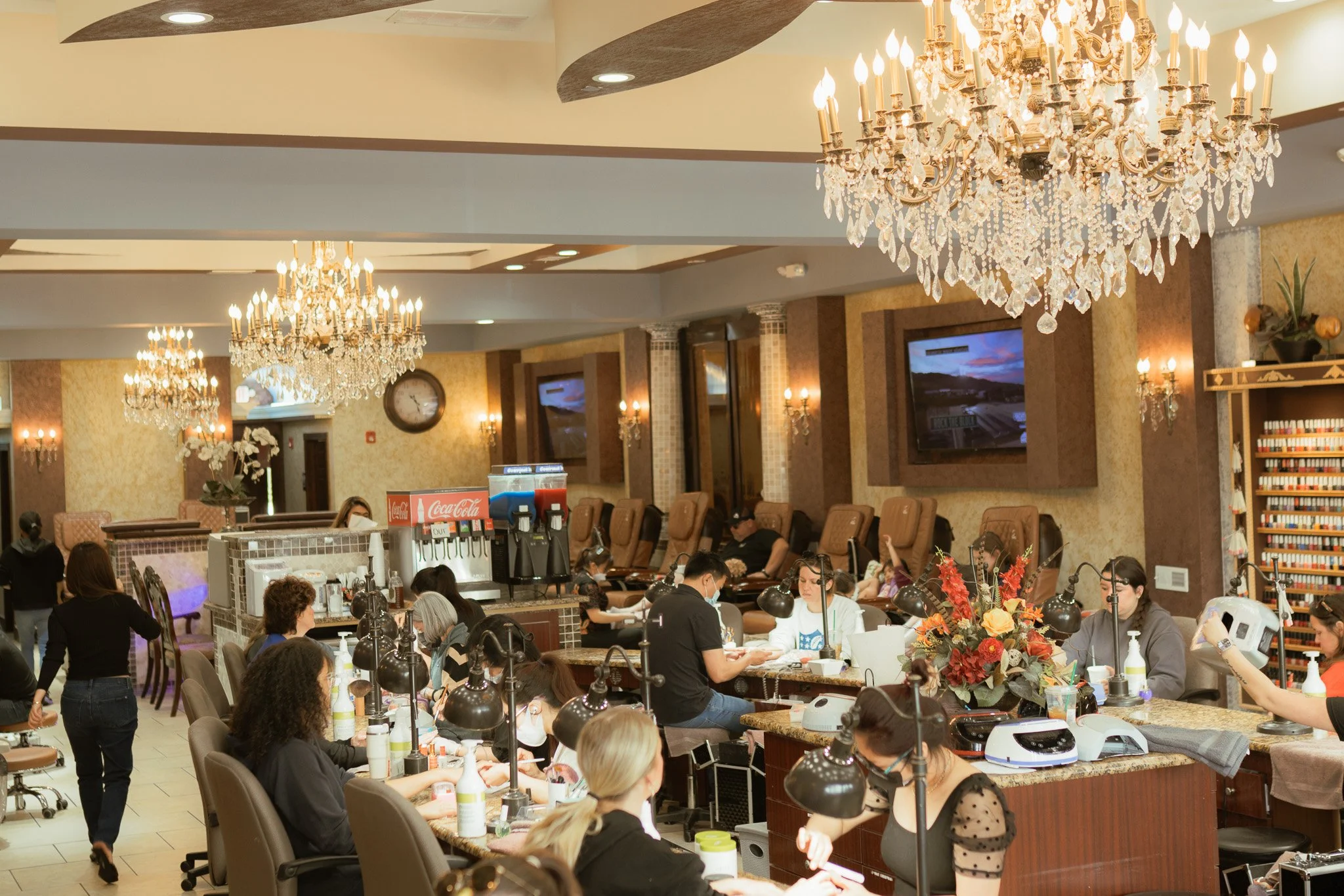 Interior of a nail salon with clients receiving manicures, several chairs for pedicures along the wall, large chandeliers hanging from the ceiling, and nail technicians working at stations.