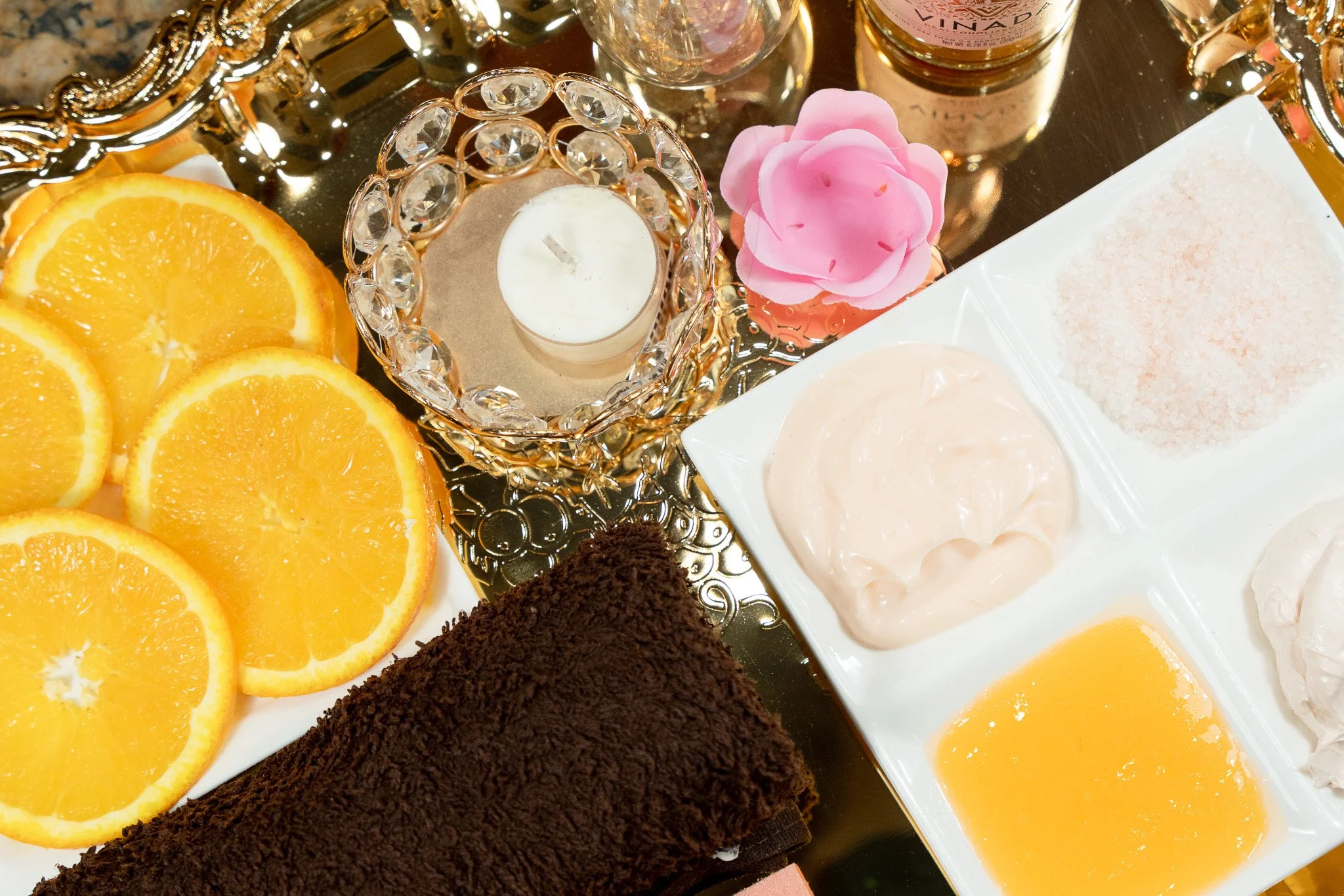 Sliced oranges, a rolled chocolate towel, and a sectioned tray of bath products including creams and salt, on a reflective surface with a decorative candle holder and pink flower.