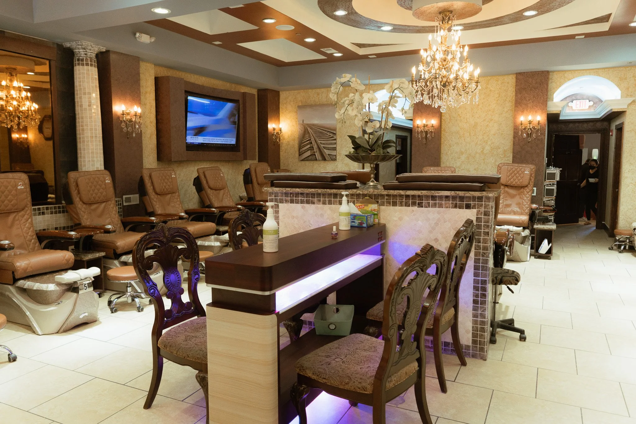 Spa reception area with massage chairs, a reception desk, a flower arrangement, a chandelier, and a wall-mounted TV.