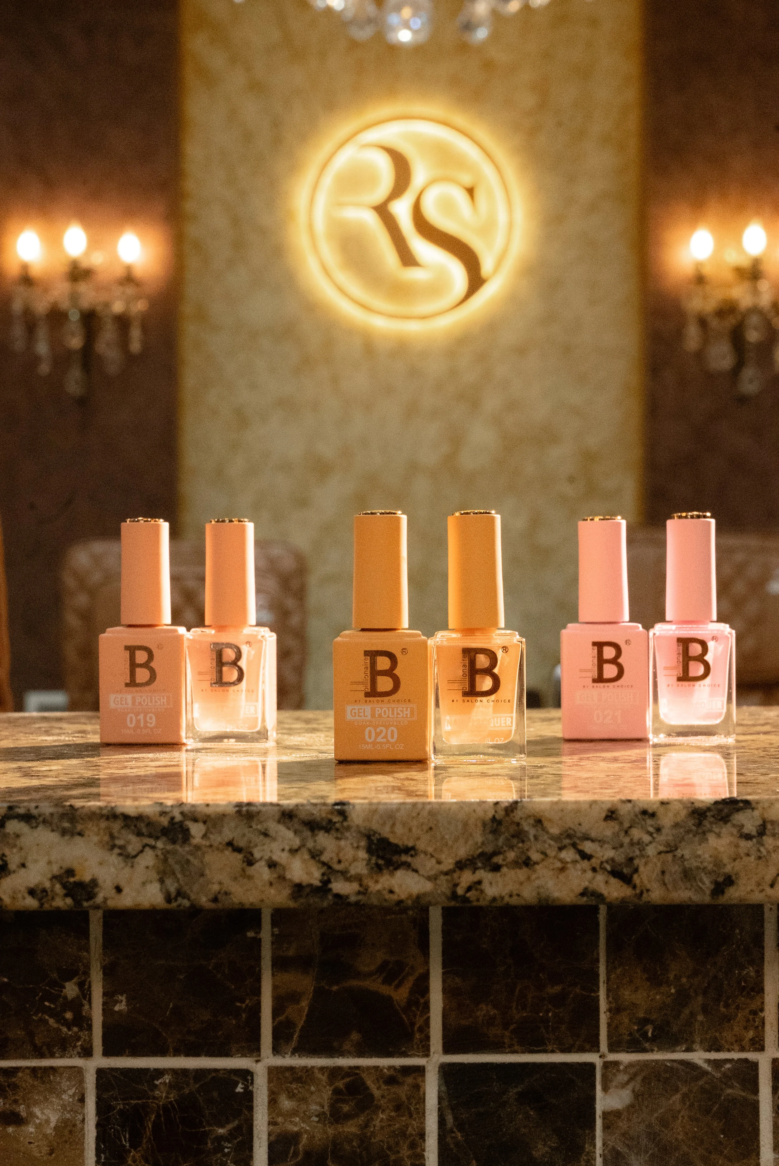 Six bottles of gel nail polish displayed on a marble countertop, with three pink bottles on the right and three beige bottles in the center, in a warmly lit room with a logo illuminated on the textured wall behind. Rock Star nails new collection