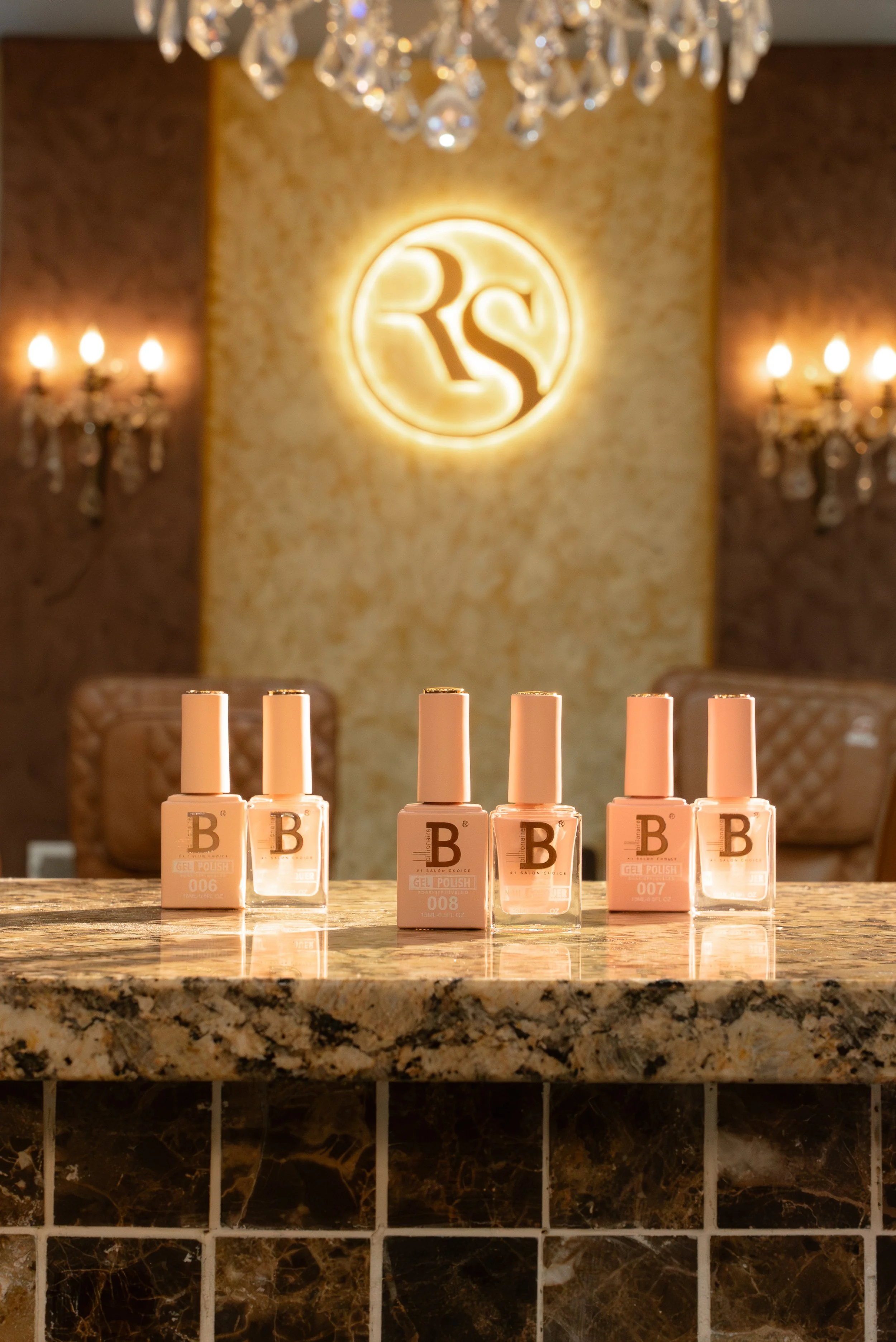 Six bottles of pink gel nail polish are displayed on a marble countertop in a luxurious salon with a chandelier and illuminated logo in the background. Book Rock star nails