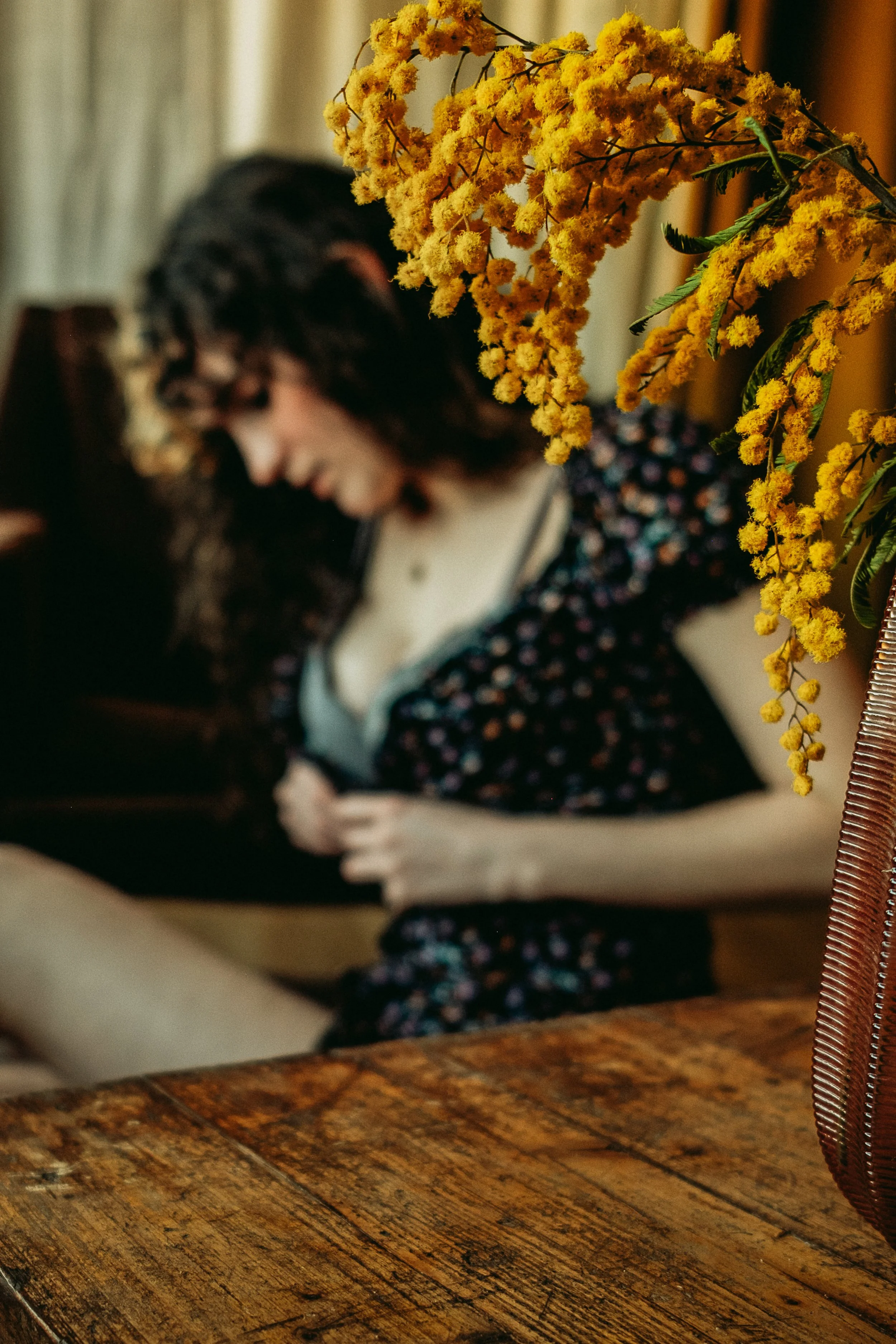 on voit du mimosa dans un salon. dérrière, floue, on distigue une femme en train de déboutenner une robe à fleur.