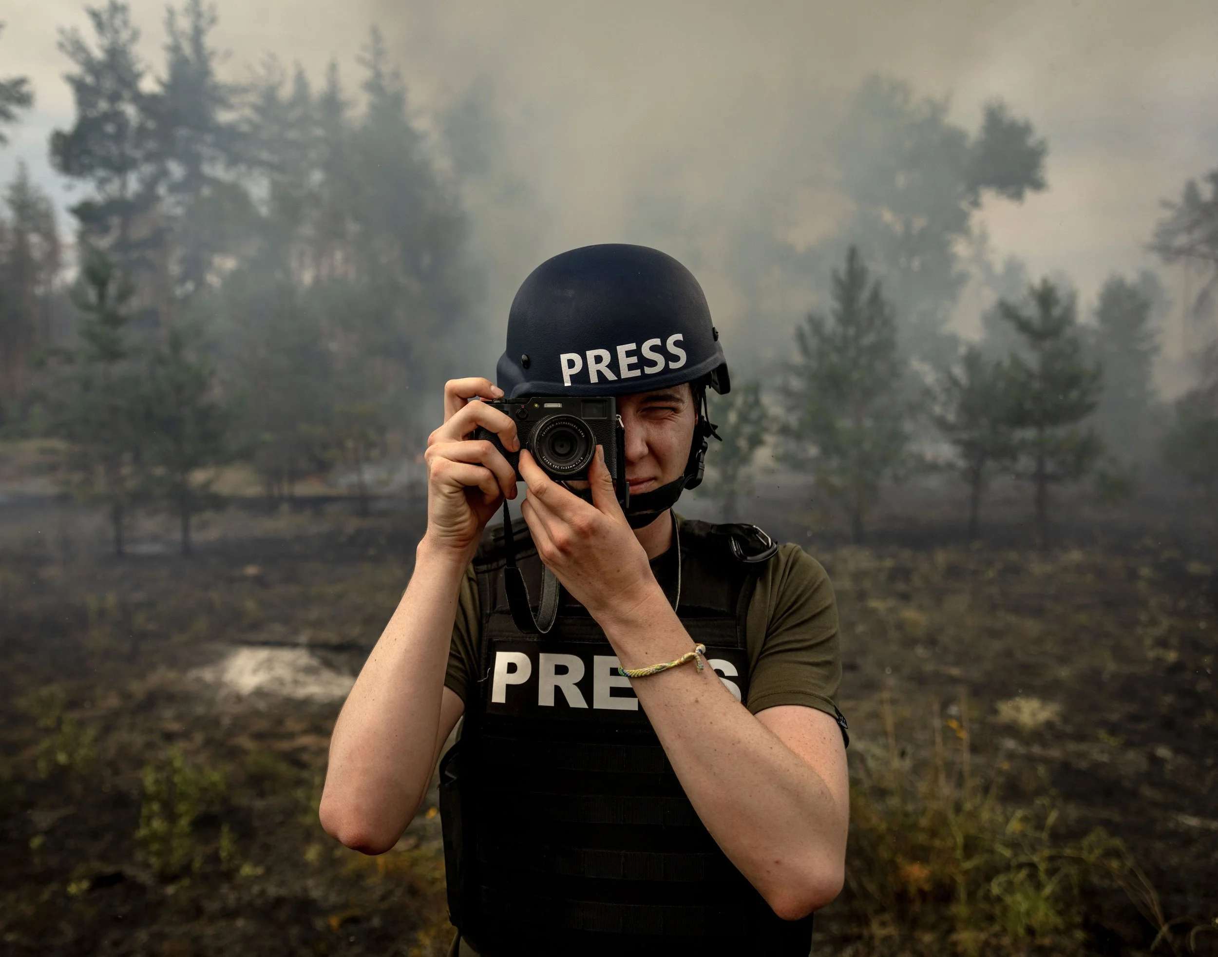 A person wearing a press helmet and vest, holding a camera, taking a photo during a wildfire with smoke and trees in the background.