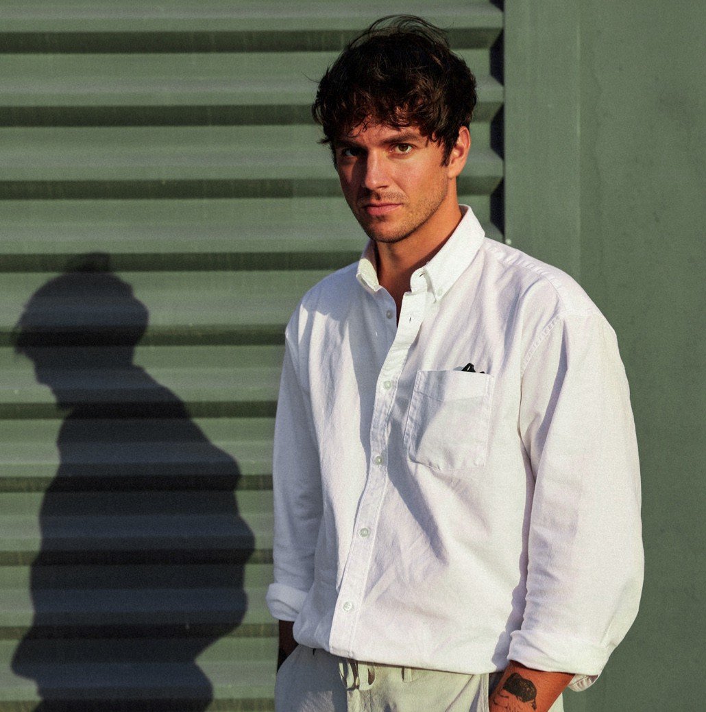 A young man with dark, curly hair, wearing a white button-up shirt with rolled-up sleeves, standing outdoors against a green wall with horizontal siding. The lighting casts a shadow of his profile on the wall.