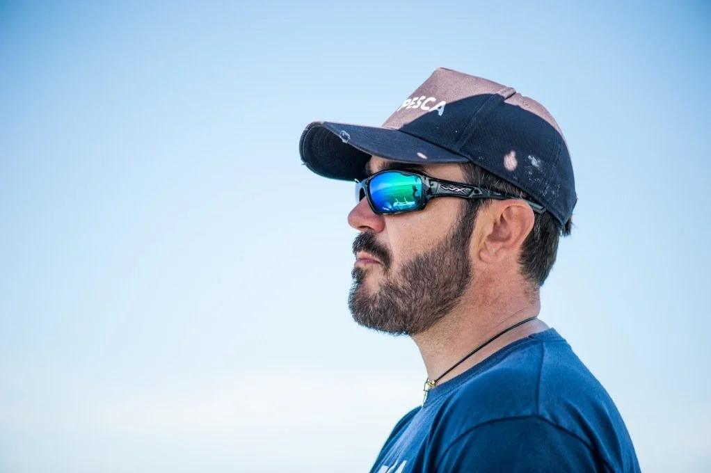 Side profile of a man wearing sunglasses, a baseball cap, and a blue T-shirt, against a clear blue sky.