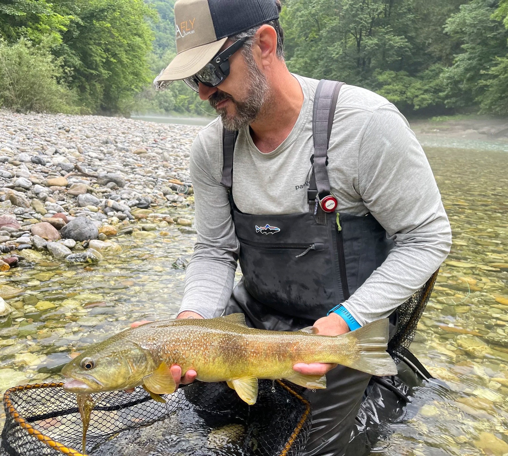 A man wearing sunglasses, a hat, and fishing gear kneels in a river, holding a large fish over a net. The riverbank with pebbles and green trees in the background.