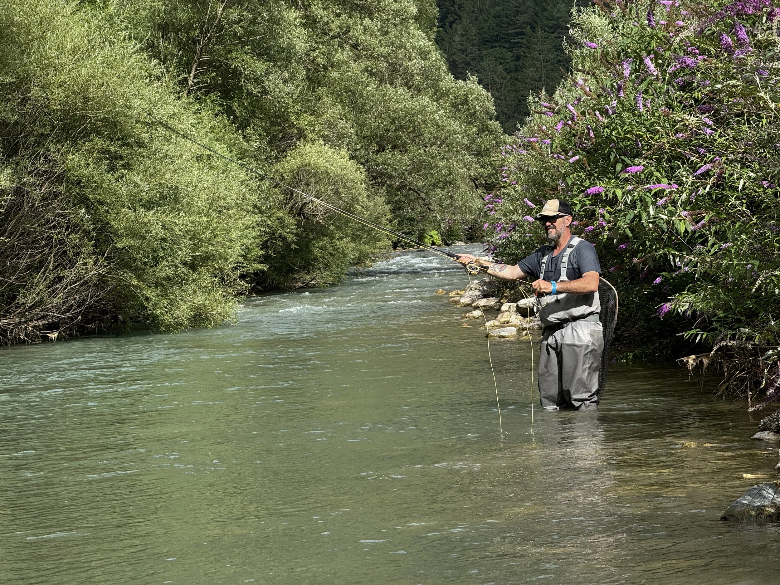 A man fishing in a river with lush green trees and bushes on the banks, and purple flowering plants nearby.