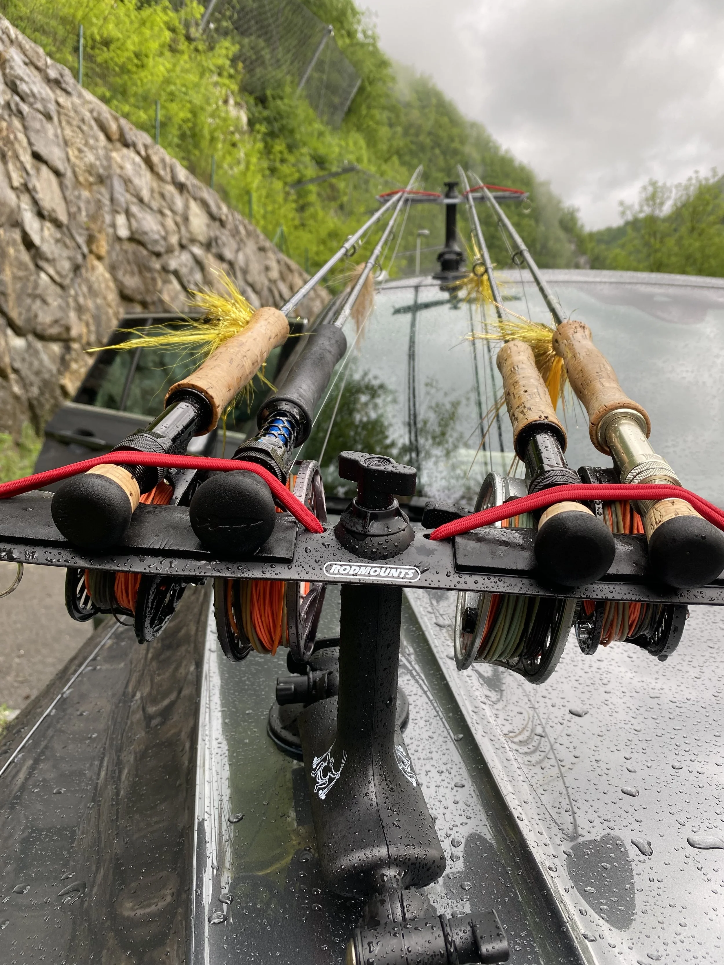 Fishing tackle and gear on a fallen log by a river, including an orange Megafly tackle box, a tan Megafly cap, and four yellow fishing lures with long, hair-like tails.