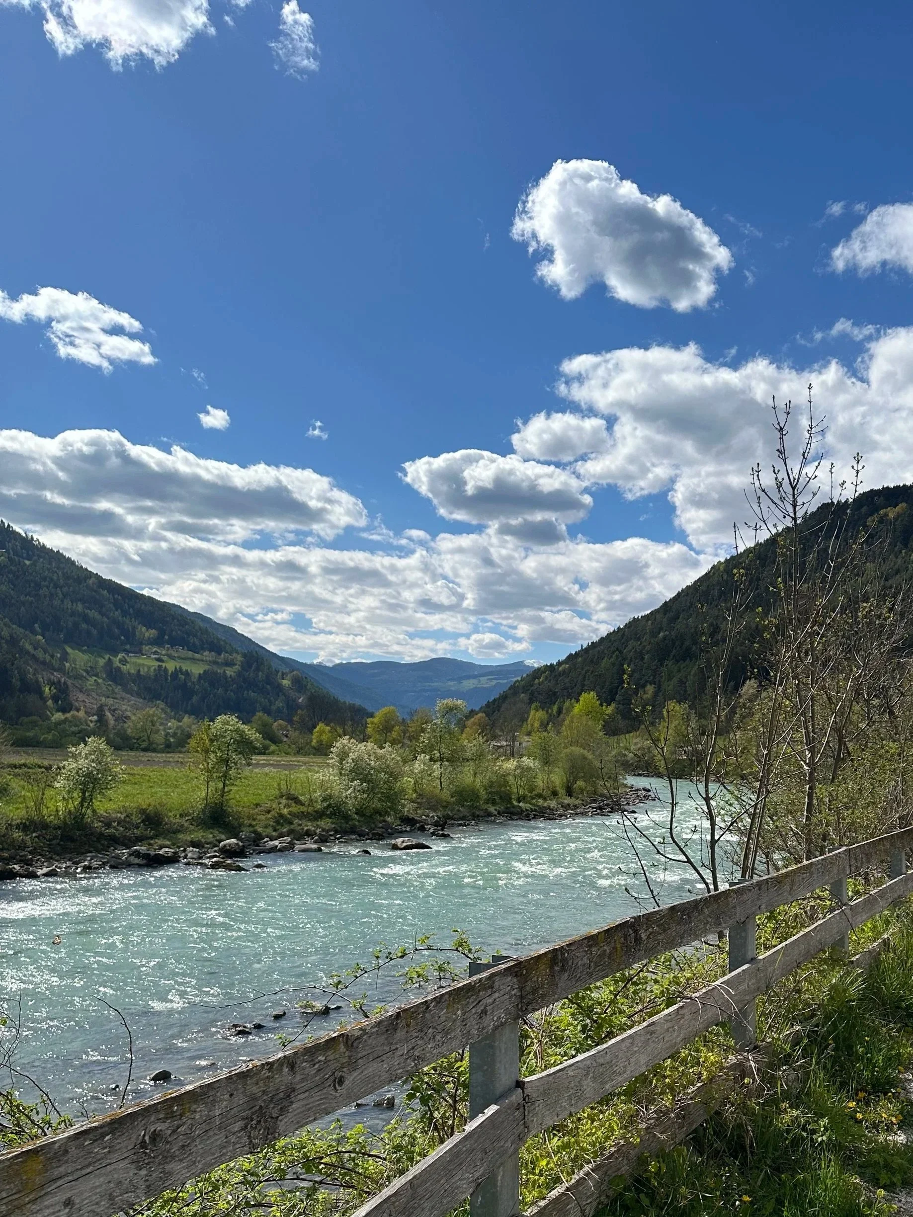 A scenic river flows through a valley bordered by green mountains with trees, under a partly cloudy blue sky.