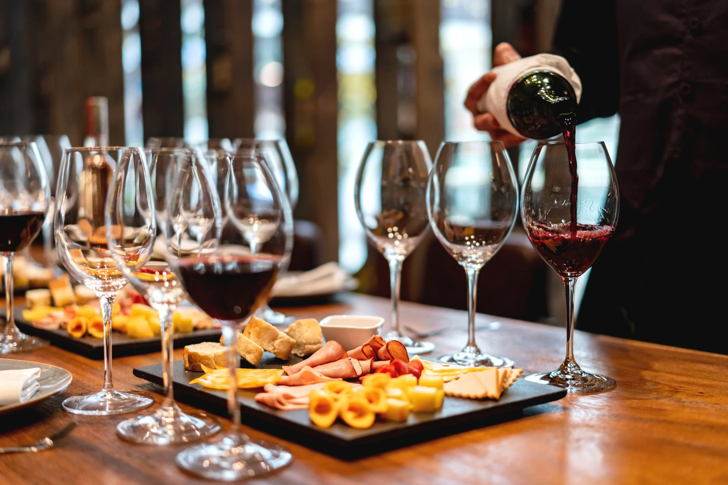 A person pouring red wine into a glass on a wooden table with multiple wine glasses and a cheese and charcuterie platter.