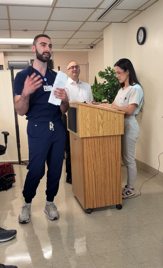 Bearded male medical student in dark blue scrubs speaks, female medical student takes notes, teacher in the background