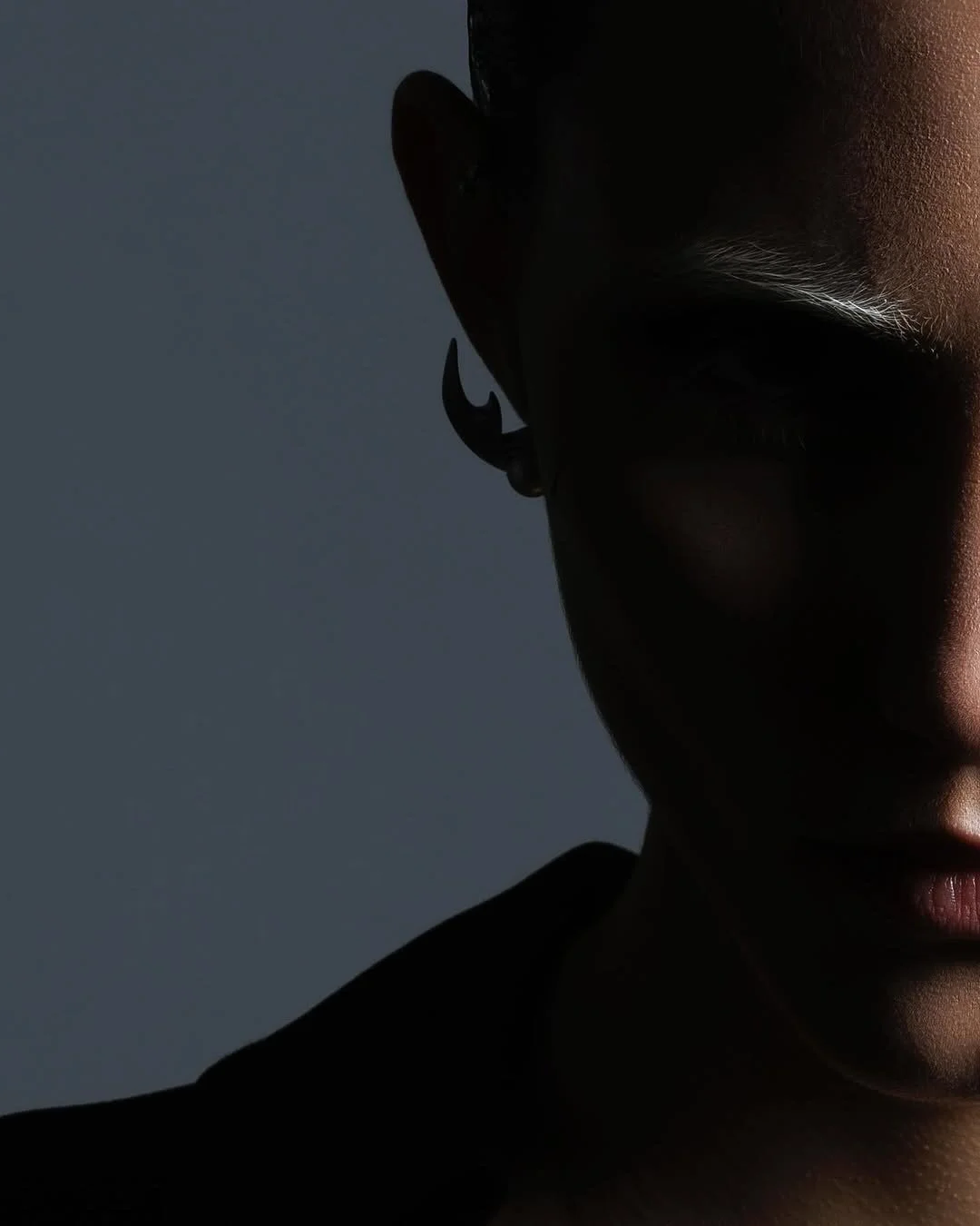 Close-up of the right side of a person's face with dark skin, focusing on part of their face, ear with a hoop earring, and eyebrow, with dramatic low-key lighting creating shadows.