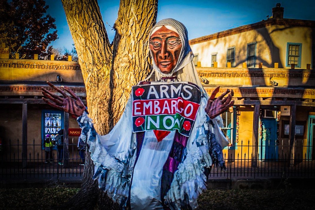 Winter Crone puppet with Arms Embargo Now banner at protest vigil for Palestinian Liberation in winter 2024