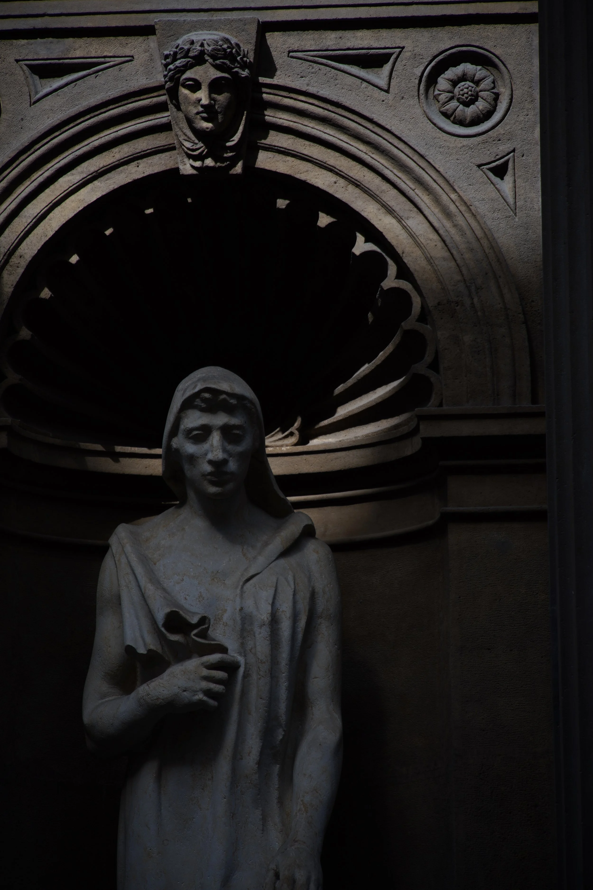 Statue of a woman in a robe holding a flower, with a carved face of a woman above and to the side, set against a decorative stone background.