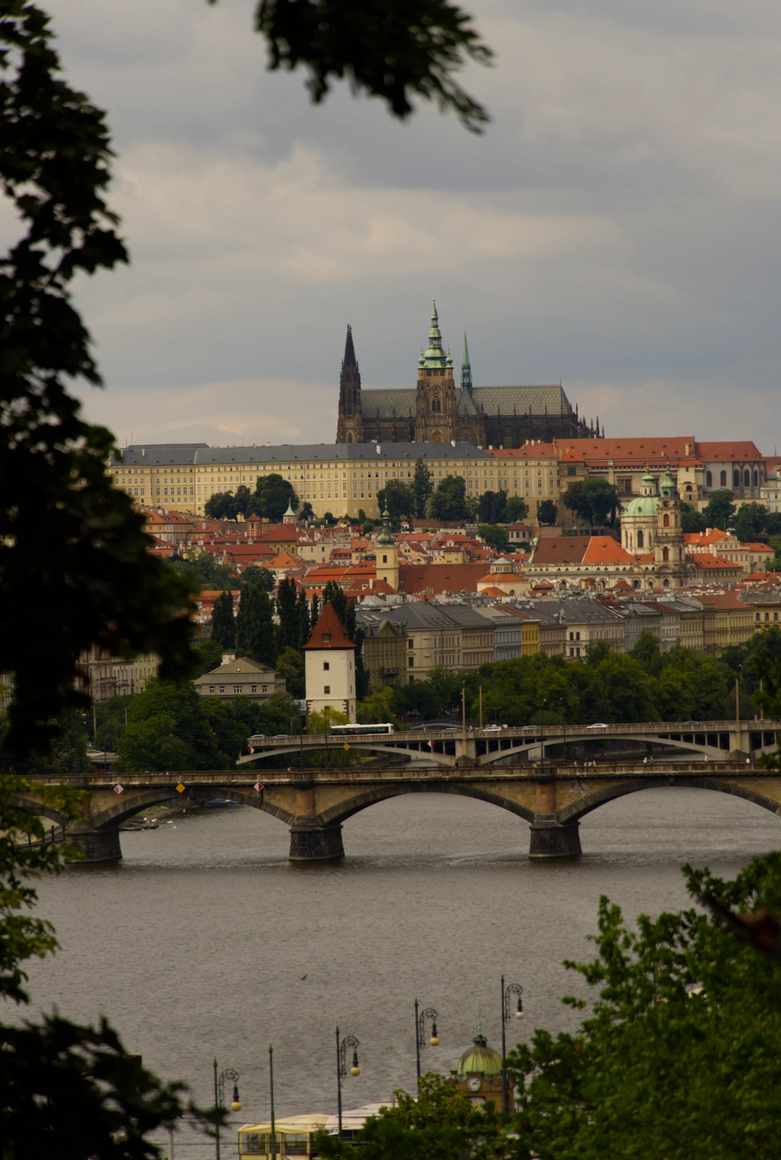 A scenic view of Prague featuring a river, a stone bridge, historic buildings, and Prague Castle with St. Vitus Cathedral in the background, under a cloudy sky.