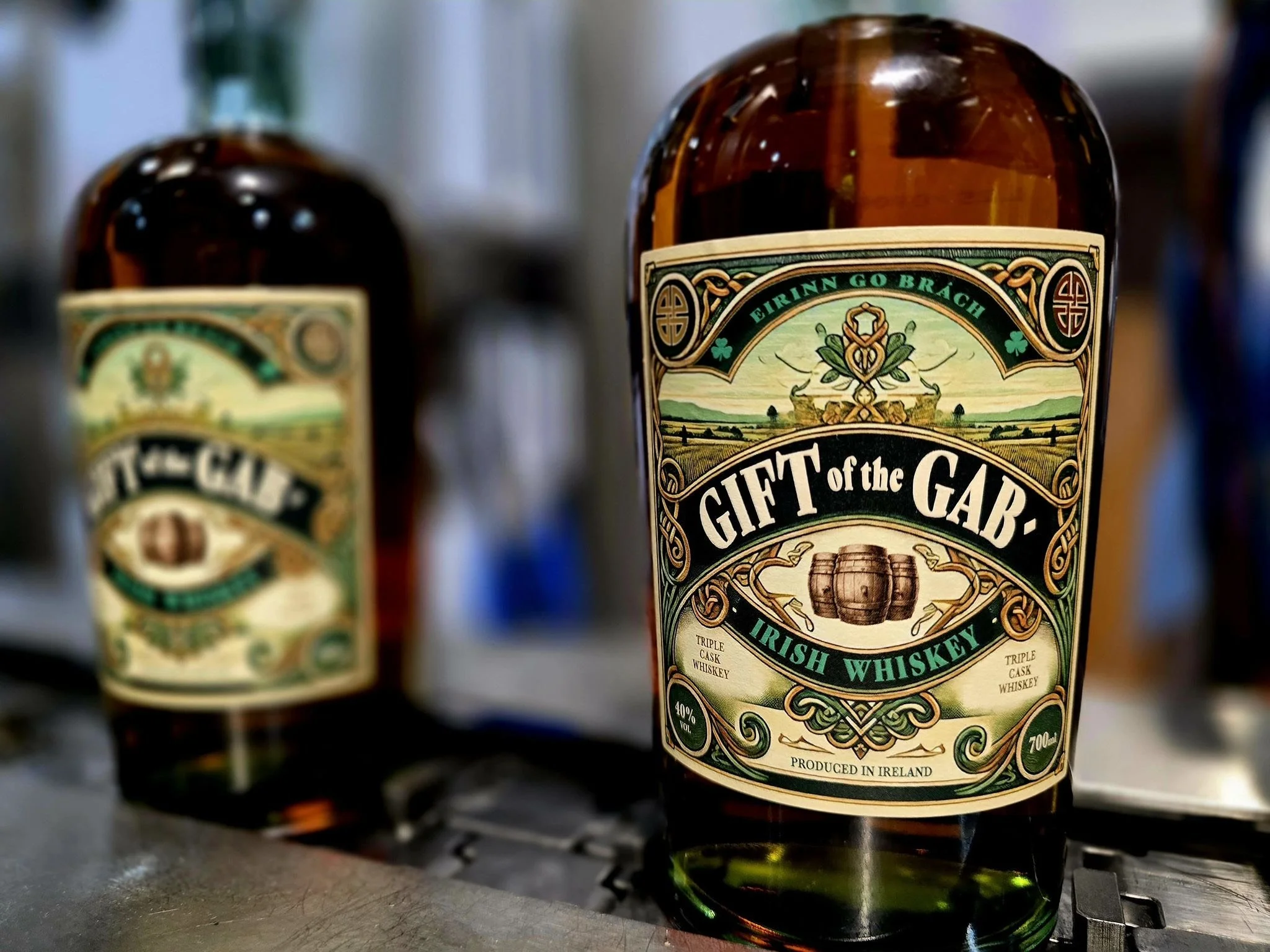 A bottle of Gift of the Gab Irish Whiskey in the foreground, with another similar whiskey bottle blurred in the background, both on a metallic surface.