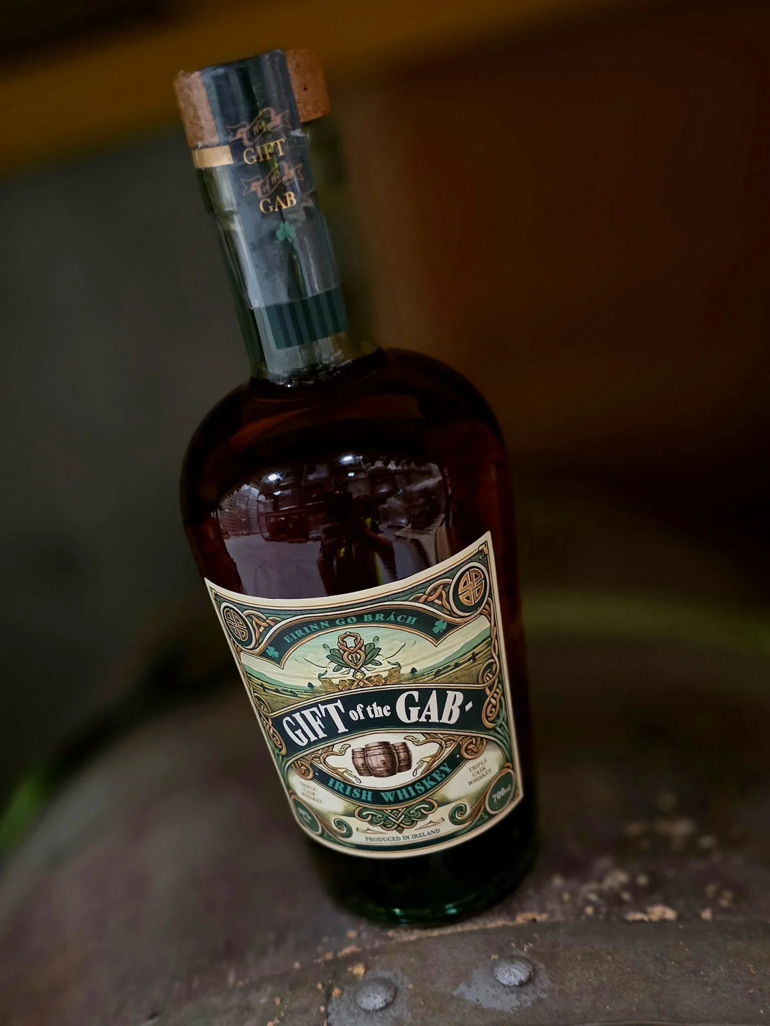 A bottle of Irish whiskey labeled 'Gift of the GAB' with a cork stopper. The label design is ornate and vintage-style, with green, gold, and white colors, and features a small barrel illustration. The bottle is placed on a wooden surface.