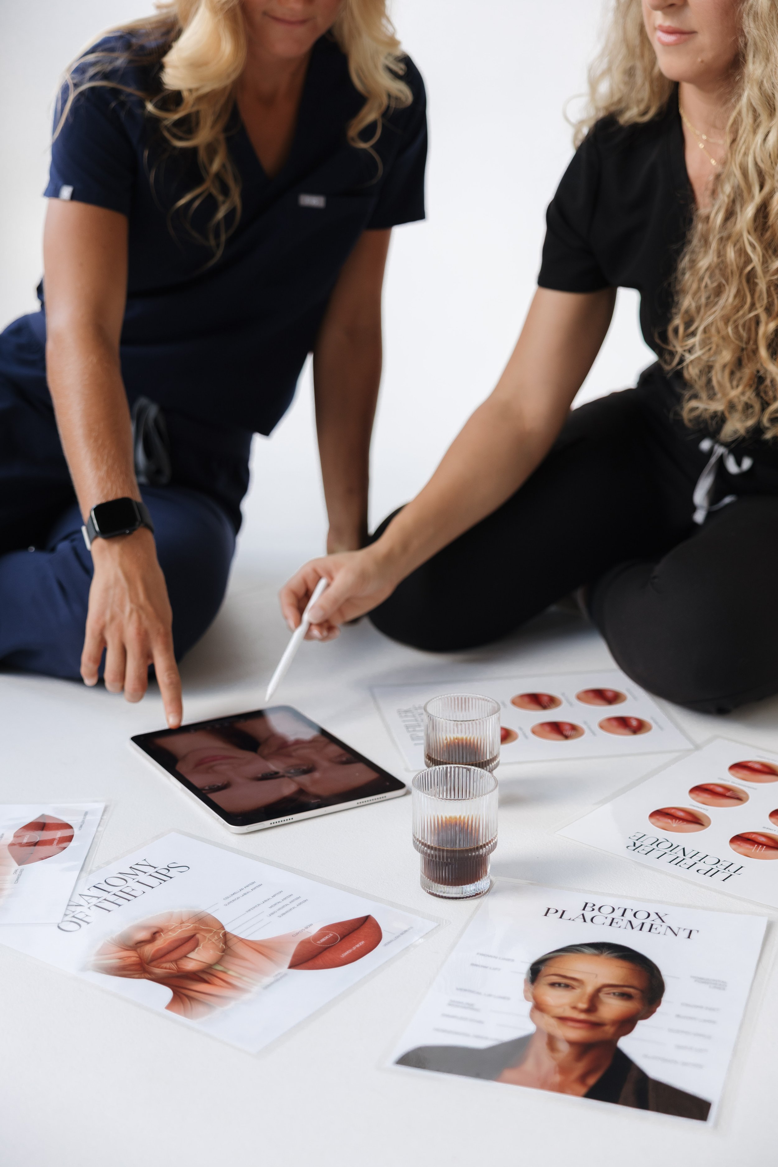 Two women are sitting at a desk covered with diagrams and information about lip and botox treatments, with glass of liquid, tablet device, and printed materials, discussing cosmetic procedures.