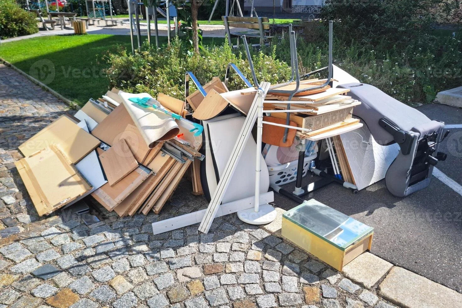 Piles of discarded furniture and household items, including wooden panels, metal stands, a lamp, a skateboard, and a glass-topped box, set on a cobblestone and asphalt sidewalk in a park.
