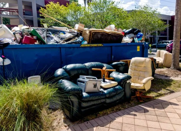 A dumpster filled with assorted trash and furniture, including couches and chairs, near a sidewalk with green plants and trees in the background.