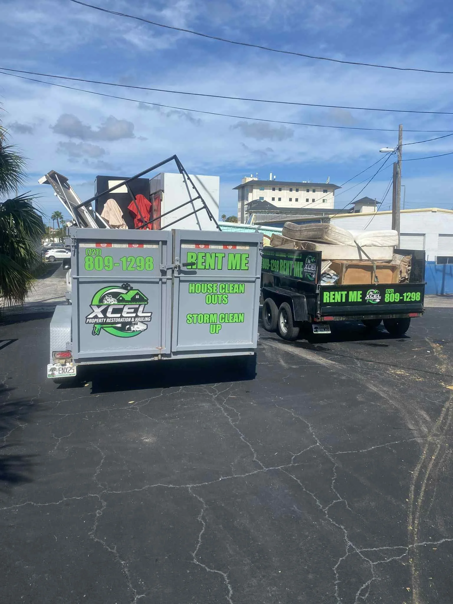 Two trucks parked in a lot with furniture loaded on them. The trucks are advertising property restoration and hauling services with the company name 'XCEL' and contact number 809-1298. The signs mention house clean outs and storm clean-up services.