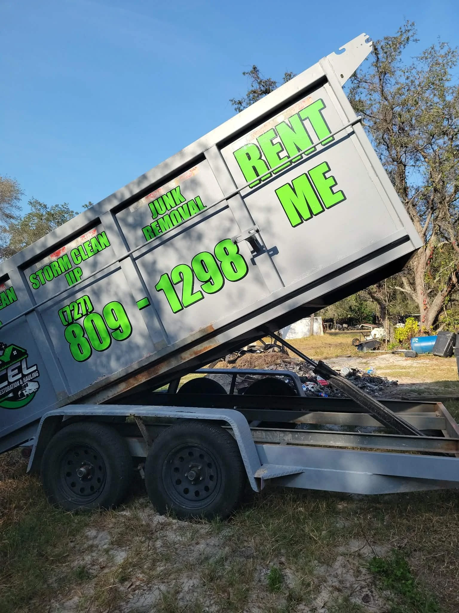 A utility trailer with a sign that reads 'Storm Clean Up, Junk Removal, Rent Me' and a phone number, parked outdoors in a grassy area with trees in the background.