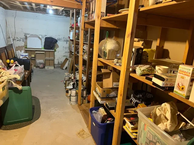 A cluttered basement storage area with wooden shelves filled with boxes, bottles, and household items. The concrete wall at the back is unfinished, and there is a green bin and a plastic container on the floor.