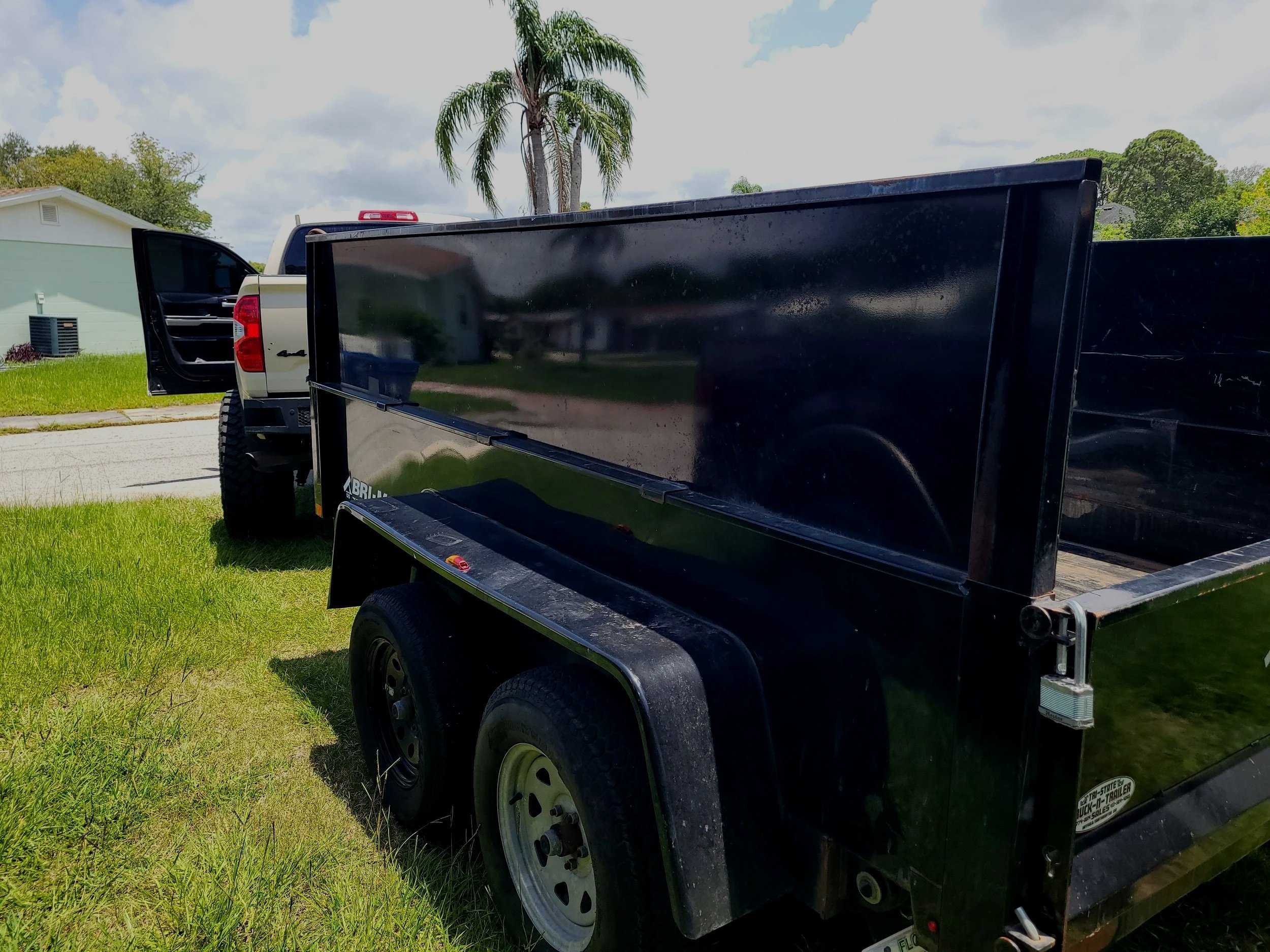 Back of a white pickup truck with an open black trailer attached, parked on a grassy area with a house, trees, and a palm tree in the background.