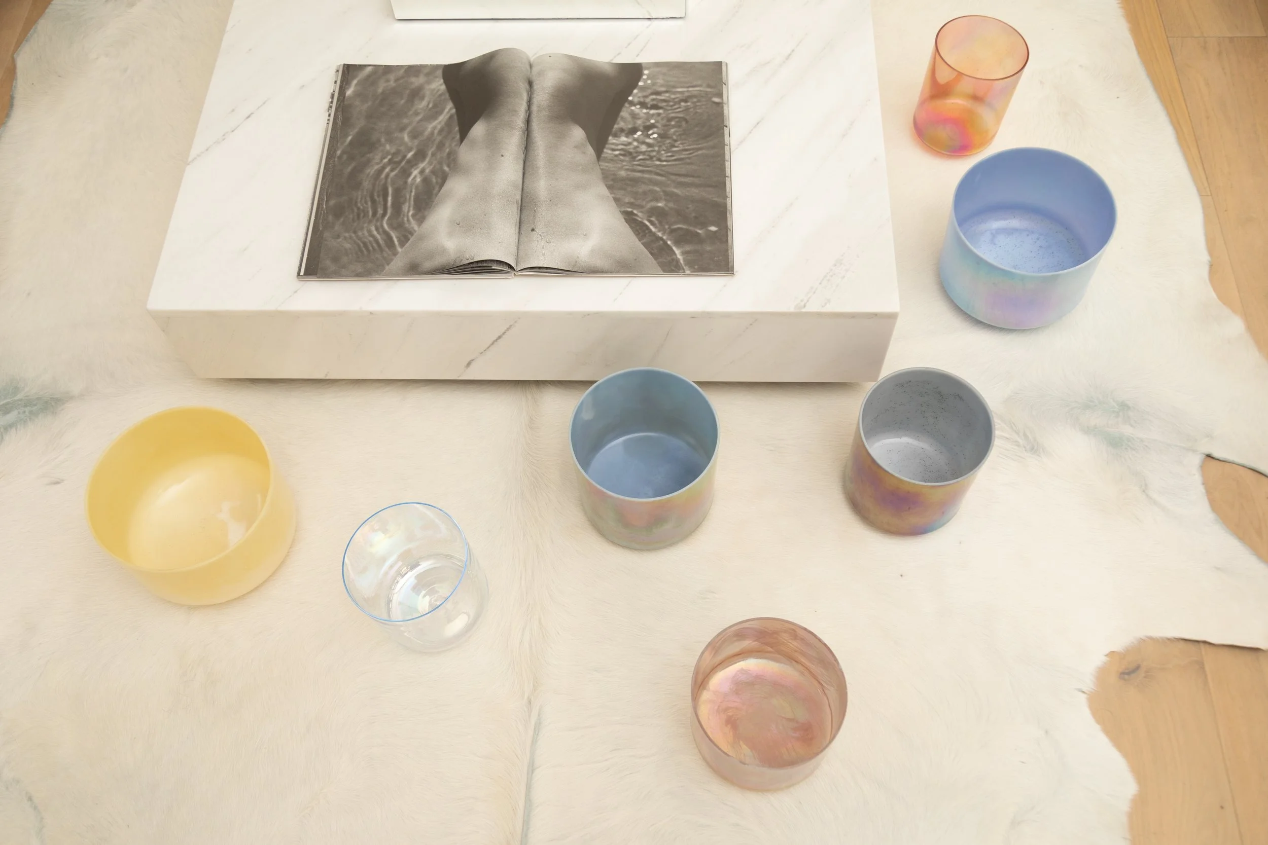 A table with a glossy white surface and a fluffy white rug underneath. On the table, there is an open magazine showing a black-and-white photo of a person's legs in water, surrounded by colorful cups of various sizes and styles, some with a pearlescent or iridescent finish, and a small transparent glass cup.