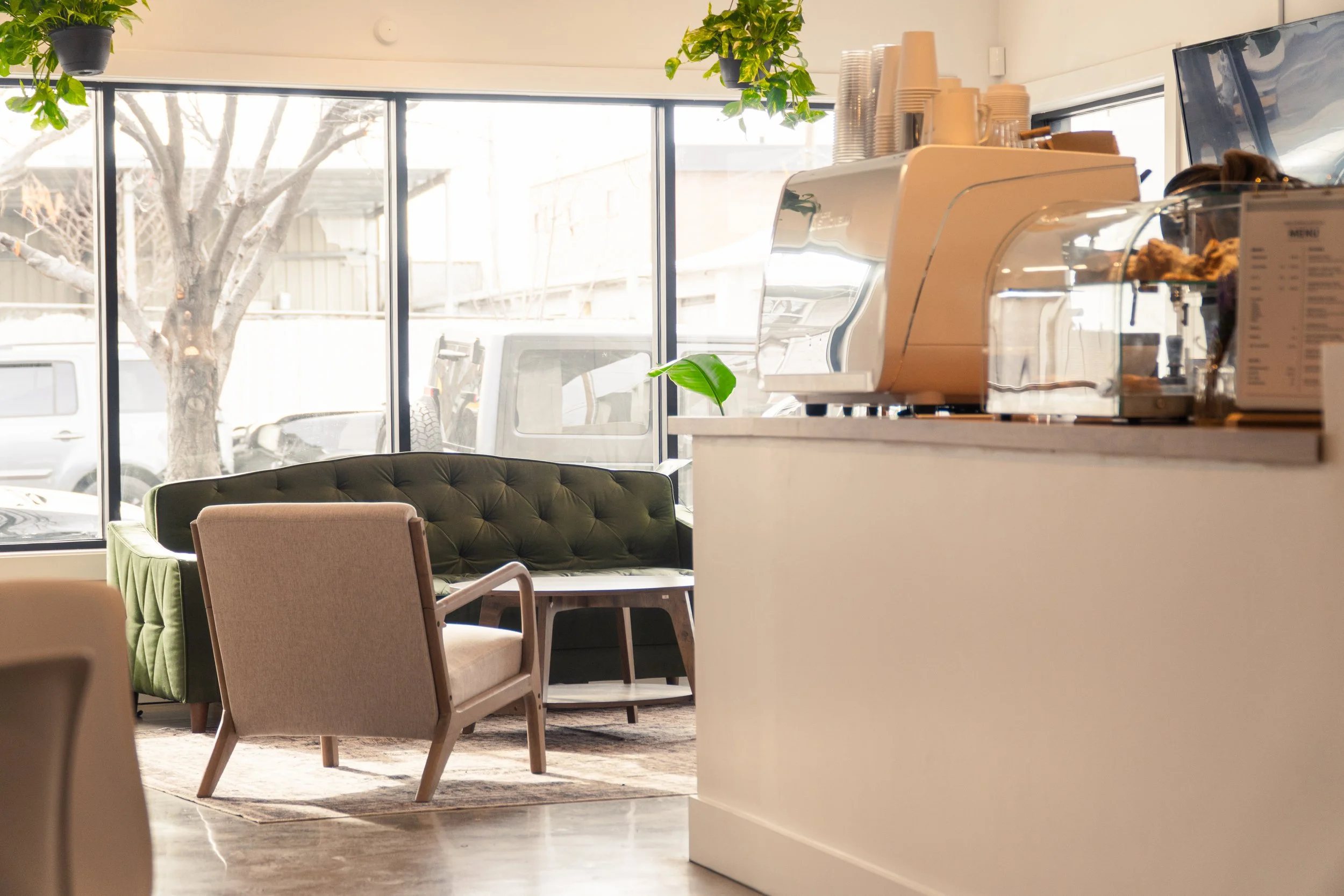Interior of a modern cafe with large windows, a green velvet sofa, a beige armchair, a coffee station, and hanging plants.