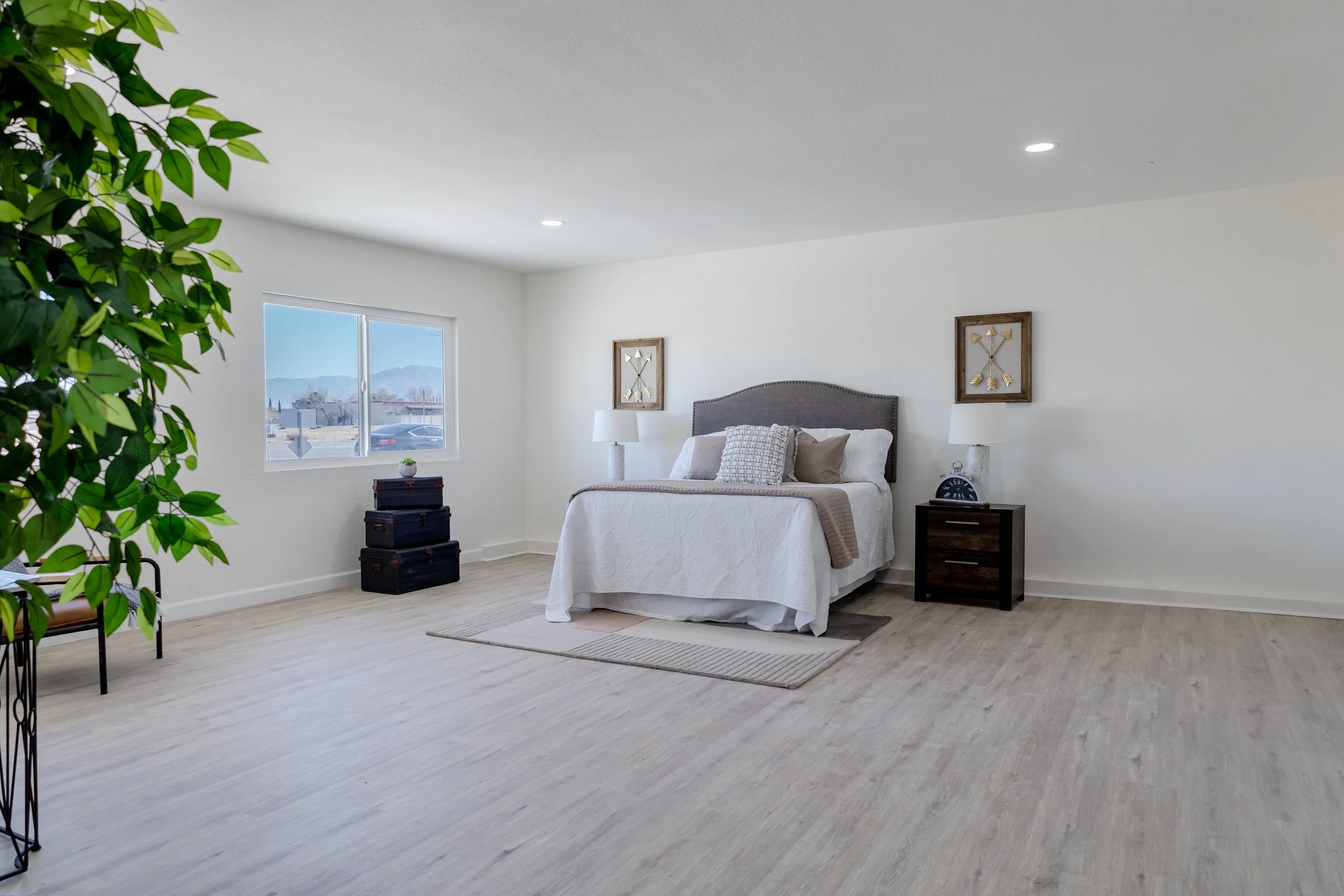 A bright, minimalistic bedroom featuring a bed with a gray headboard, white bedding, and several pillows. On each side of the bed, there are dark wooden nightstands with lamps. There is a window showing a mountain and parking lot view, and the floor