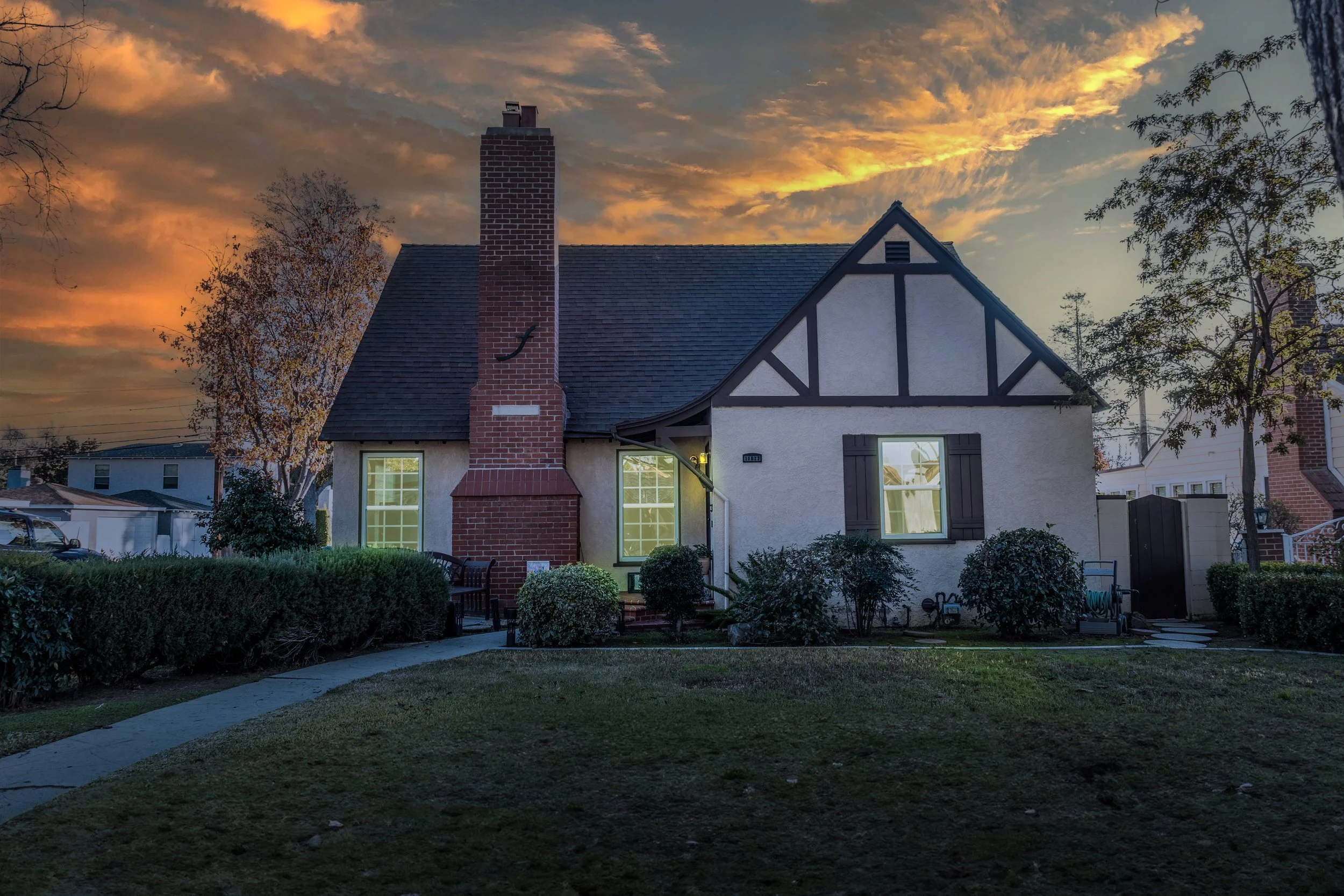 Front view of a house with a brick chimney, white stucco walls, and dark shutters, illuminated by evening light with a sunset sky in the background.