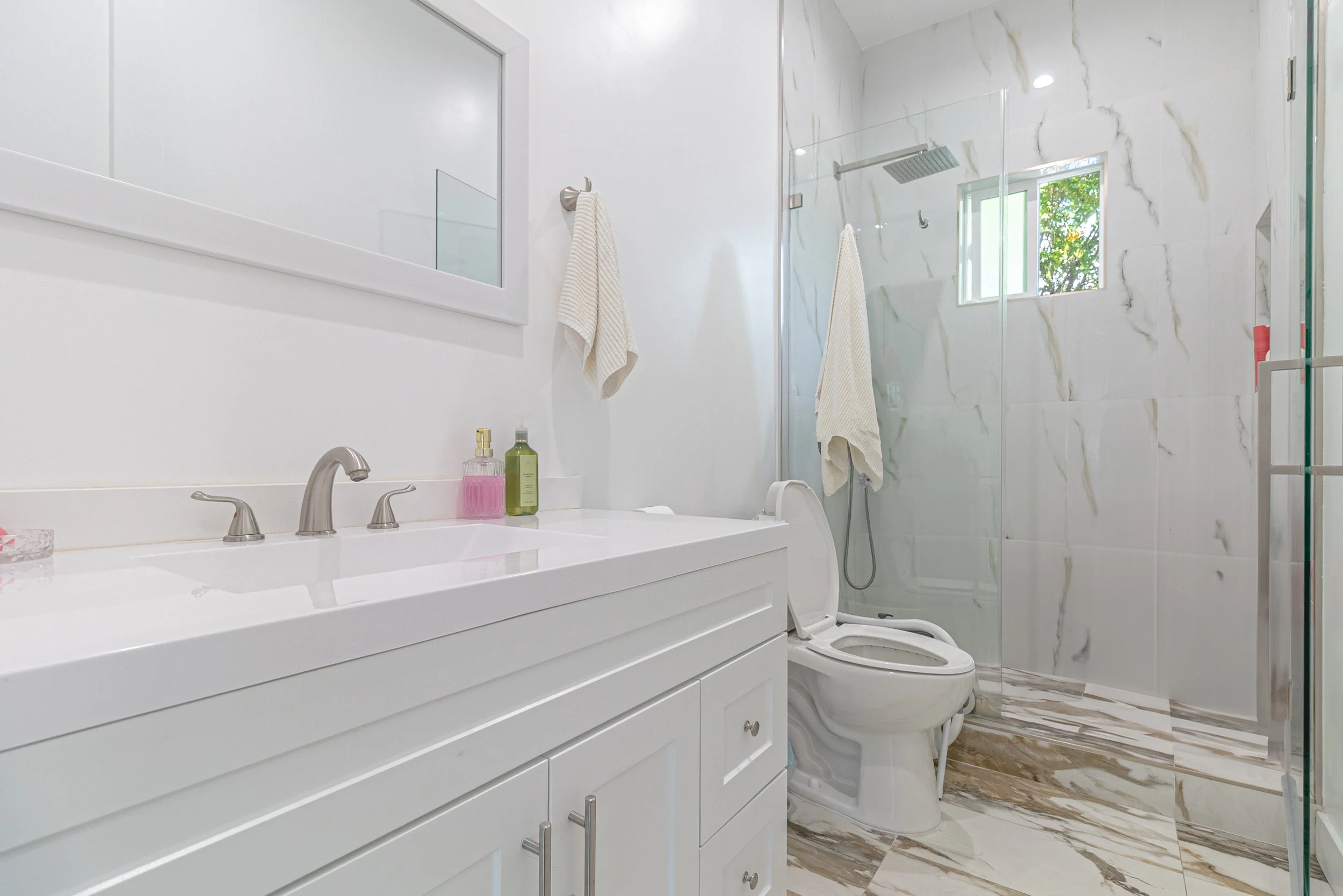 Modern bathroom with white vanity, marble floors, toilet, and glass shower enclosure with window.