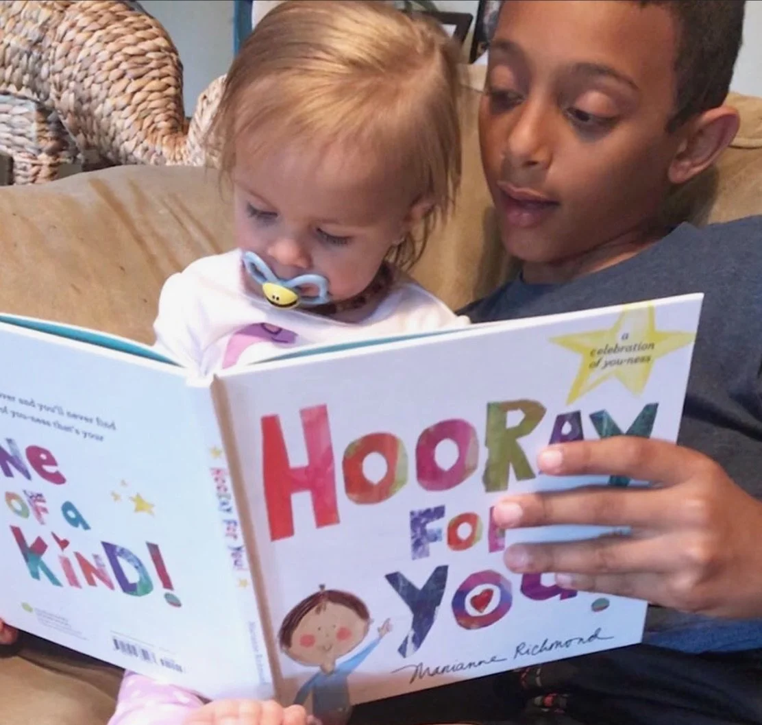 A young woman with braided hair reads a colorful children's book titled 'Hooray for You' to a small child with a pacifier. They are sitting on a beige couch with a woven chair in the background.