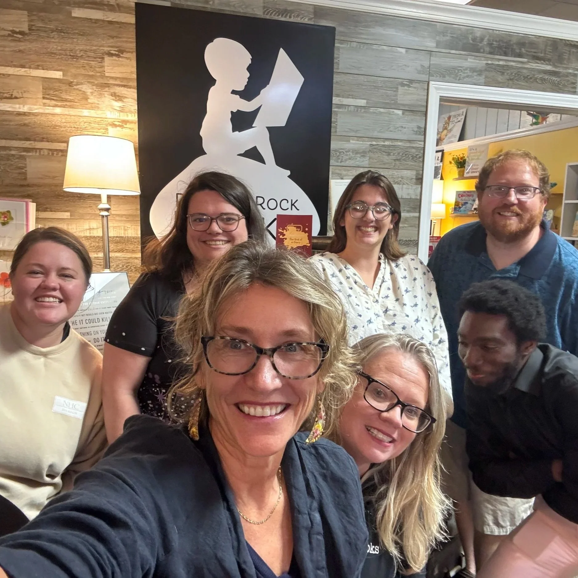 Group of eight smiling people taking a photo in front of a wooden wall with a lamp and a black poster with a silhouette of a child reading a book.