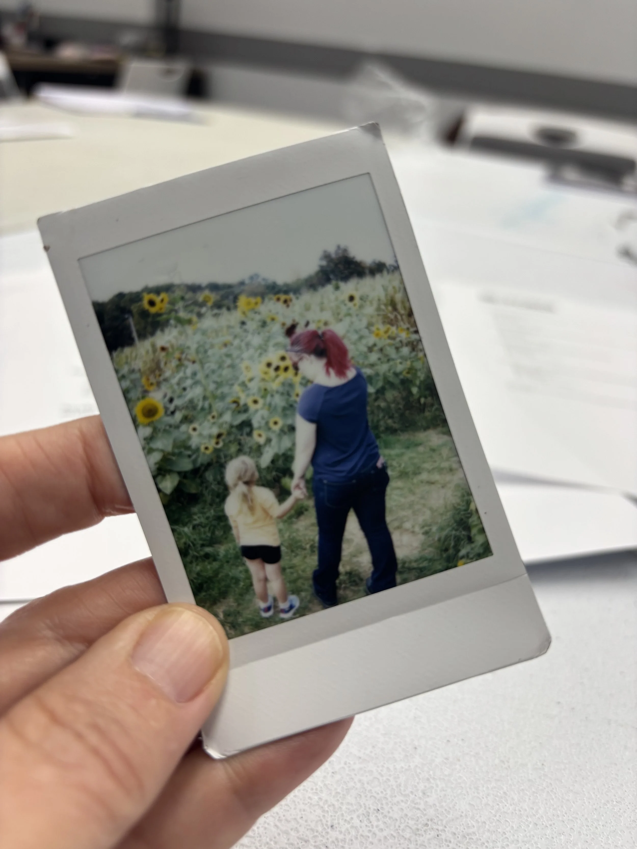 A person holding a Polaroid photo of a woman with red hair holding hands with a young girl, walking through a sunflower field.