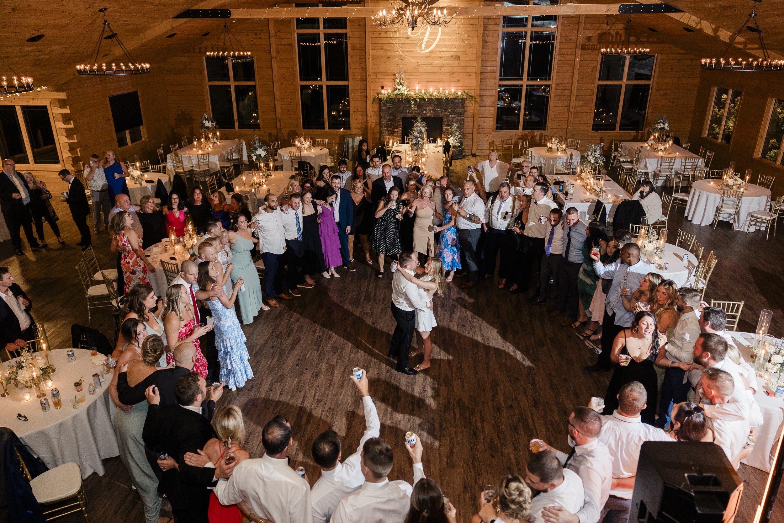 People dancing in a large wooden hall decorated for a wedding reception with round tables, floral centerpieces, and chandeliers.