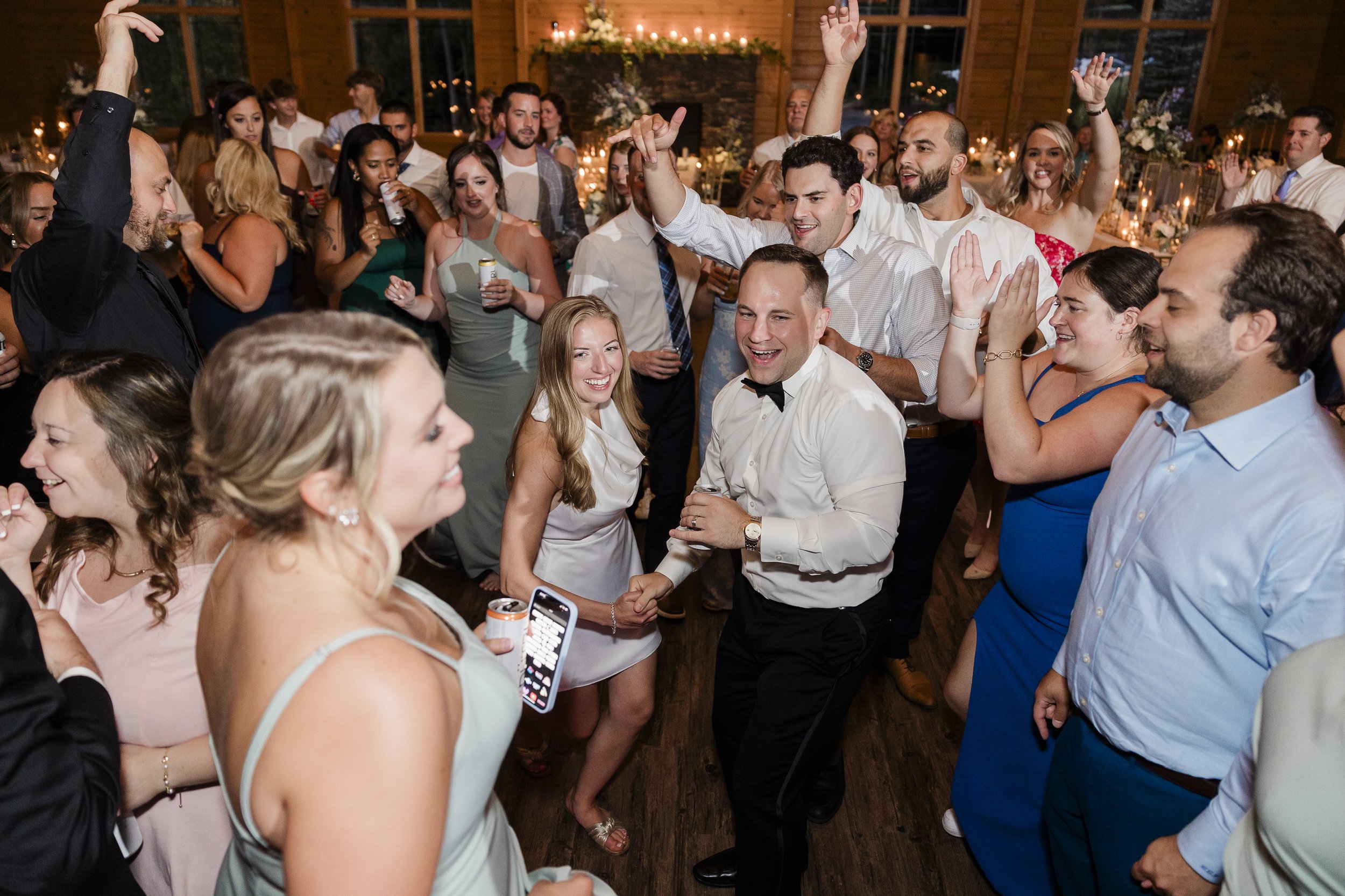 A lively group of wedding guests dancing and celebrating indoors with warm lighting and floral decorations.