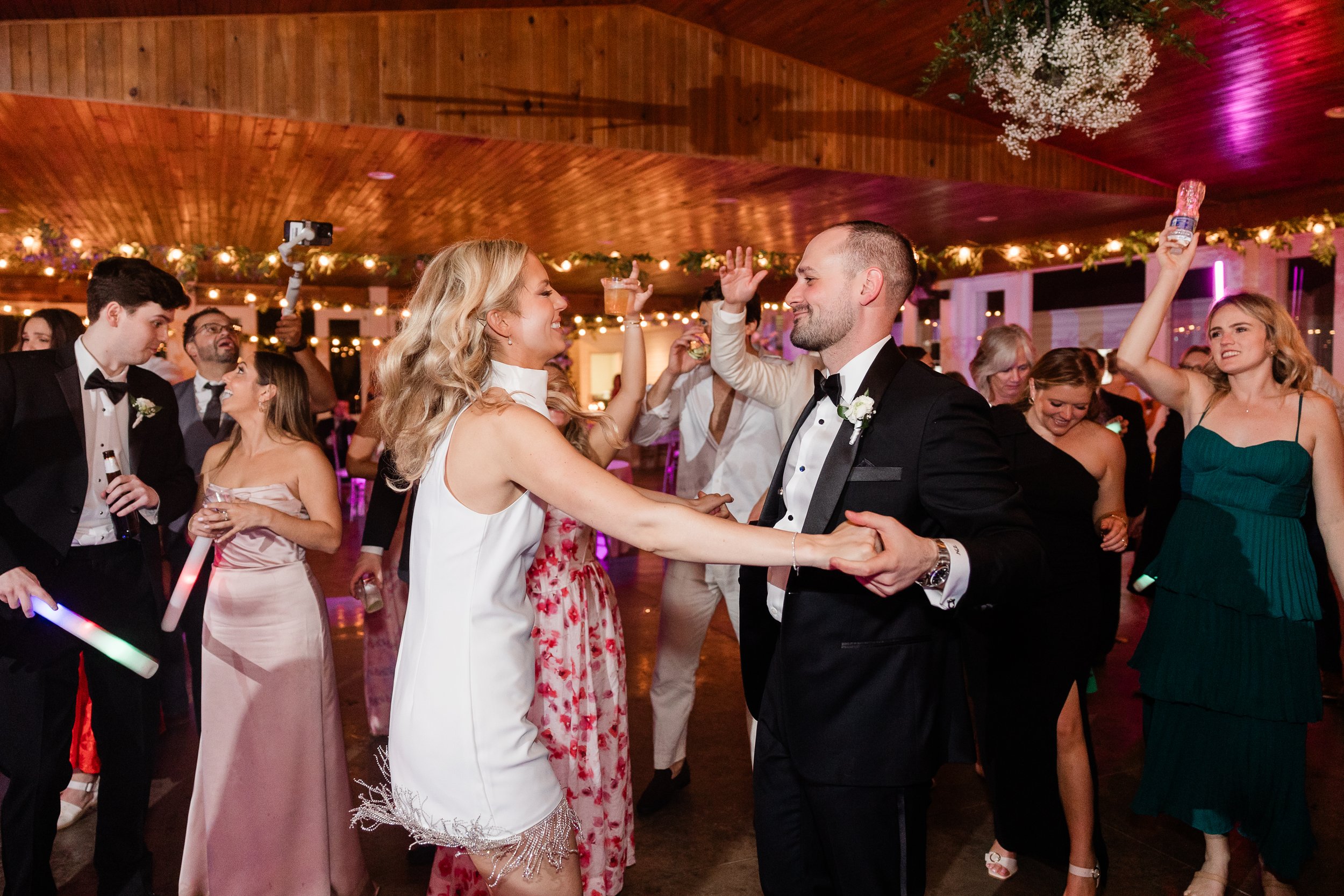 Couple dancing at a wedding reception with guests dancing and holding drinks in a decorated venue.