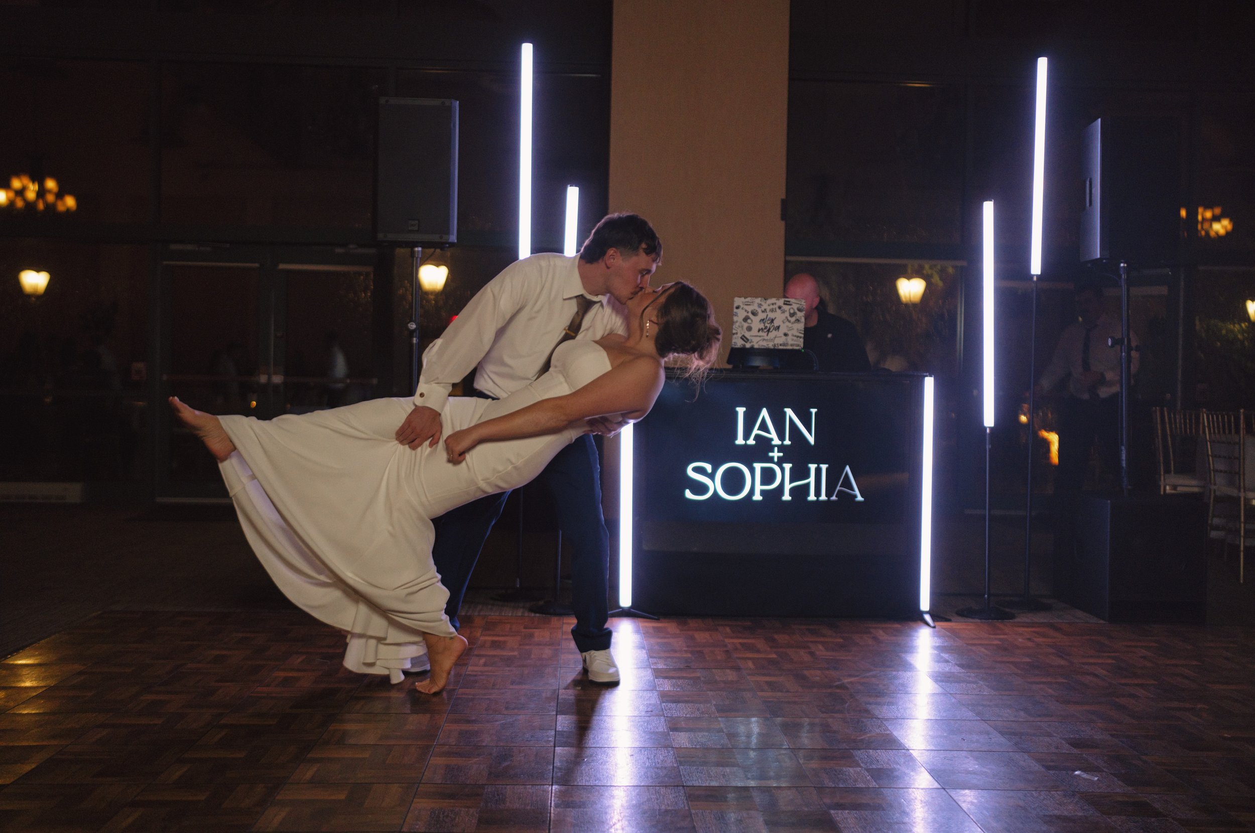 A newlywed couple sharing a dance, with the groom dipping the bride, at their wedding reception with a DJ booth in the background displaying the names 'Ian + Sophia'.