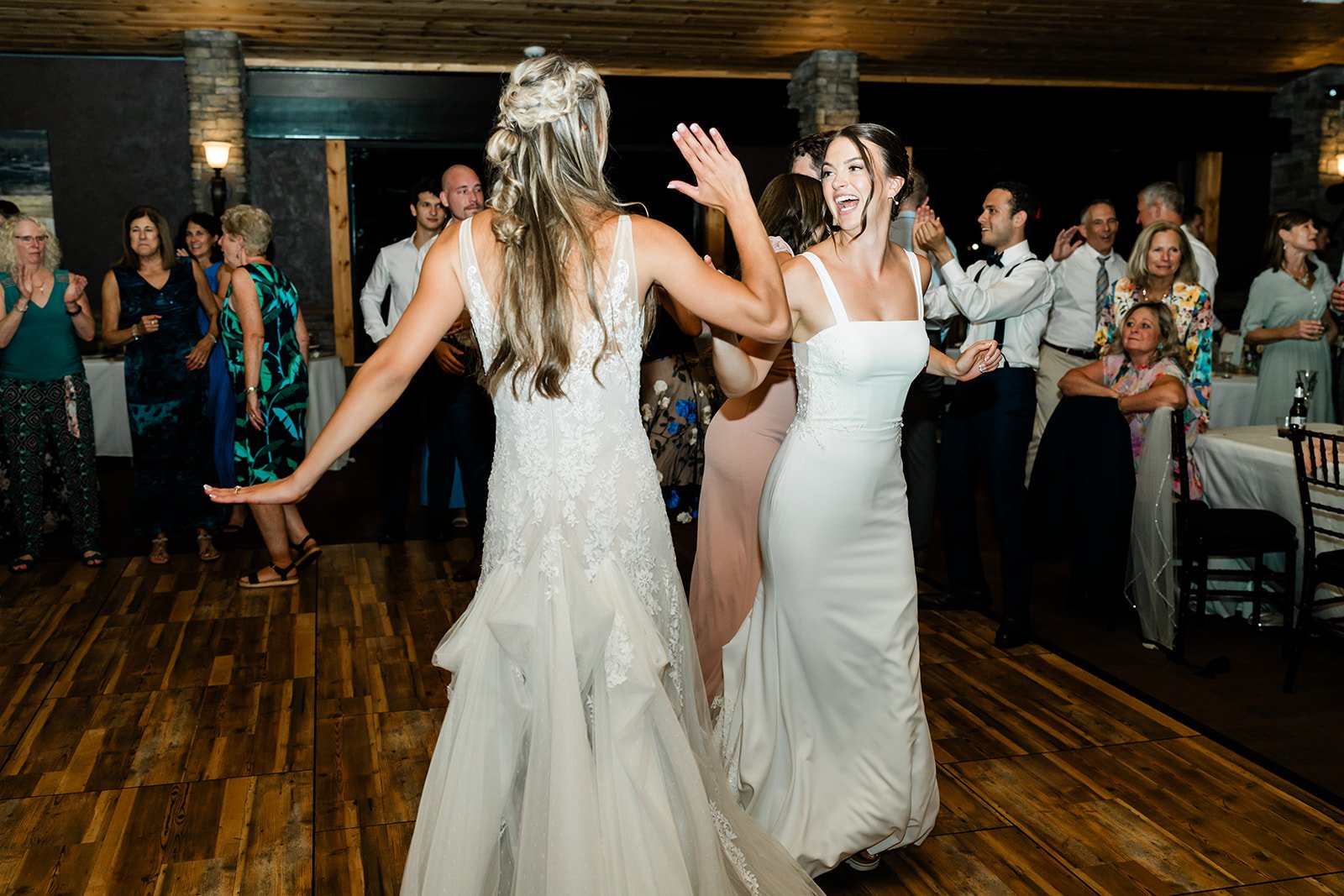 Two women in wedding dresses dancing and celebrating at a wedding reception with guests in the background.
