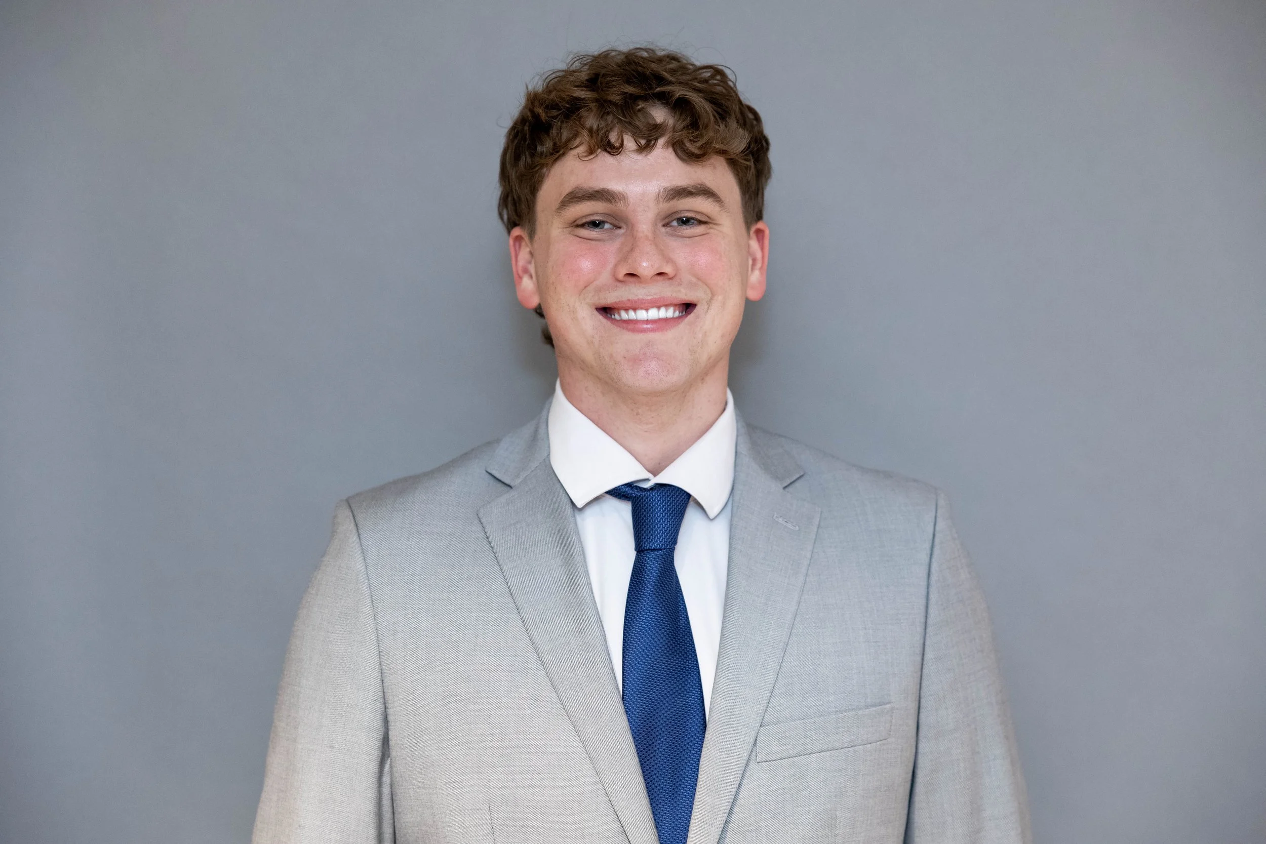A young man with curly brown hair, smiling, wearing a light gray suit jacket, white shirt, and blue tie, standing against a plain gray background.