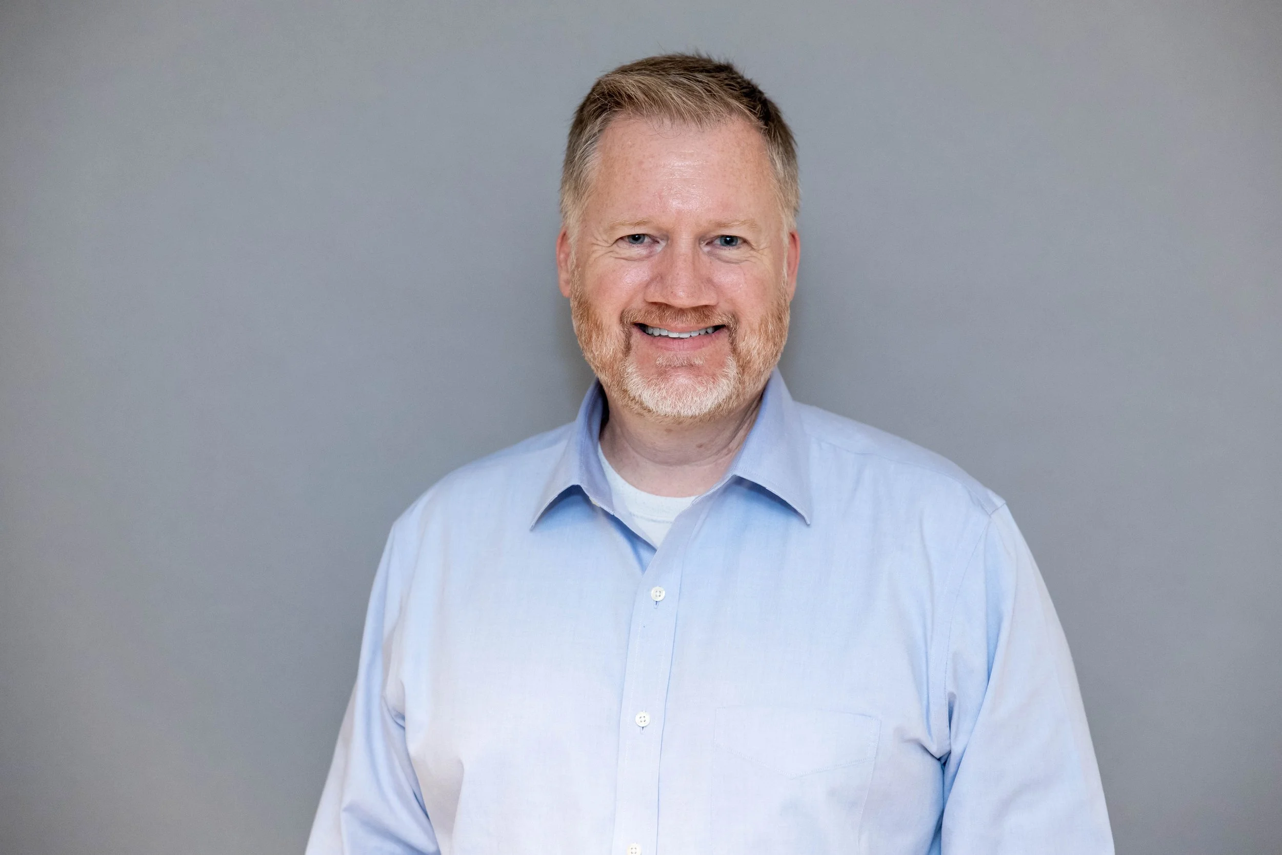A smiling middle-aged man with light-colored hair and beard wearing a light blue dress shirt, standing against a gray background.