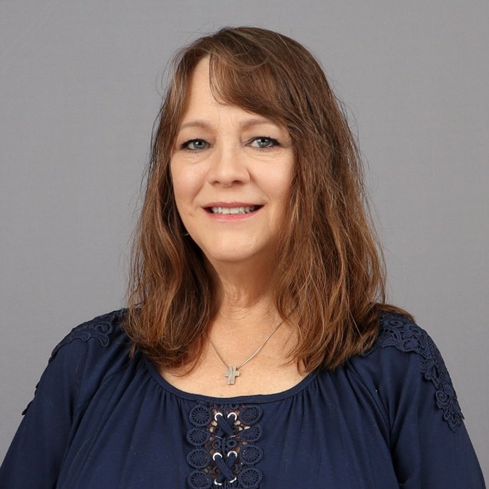 A woman with shoulder-length brown hair smiling, wearing a navy blue blouse with lace details and a cross necklace, against a plain gray background.