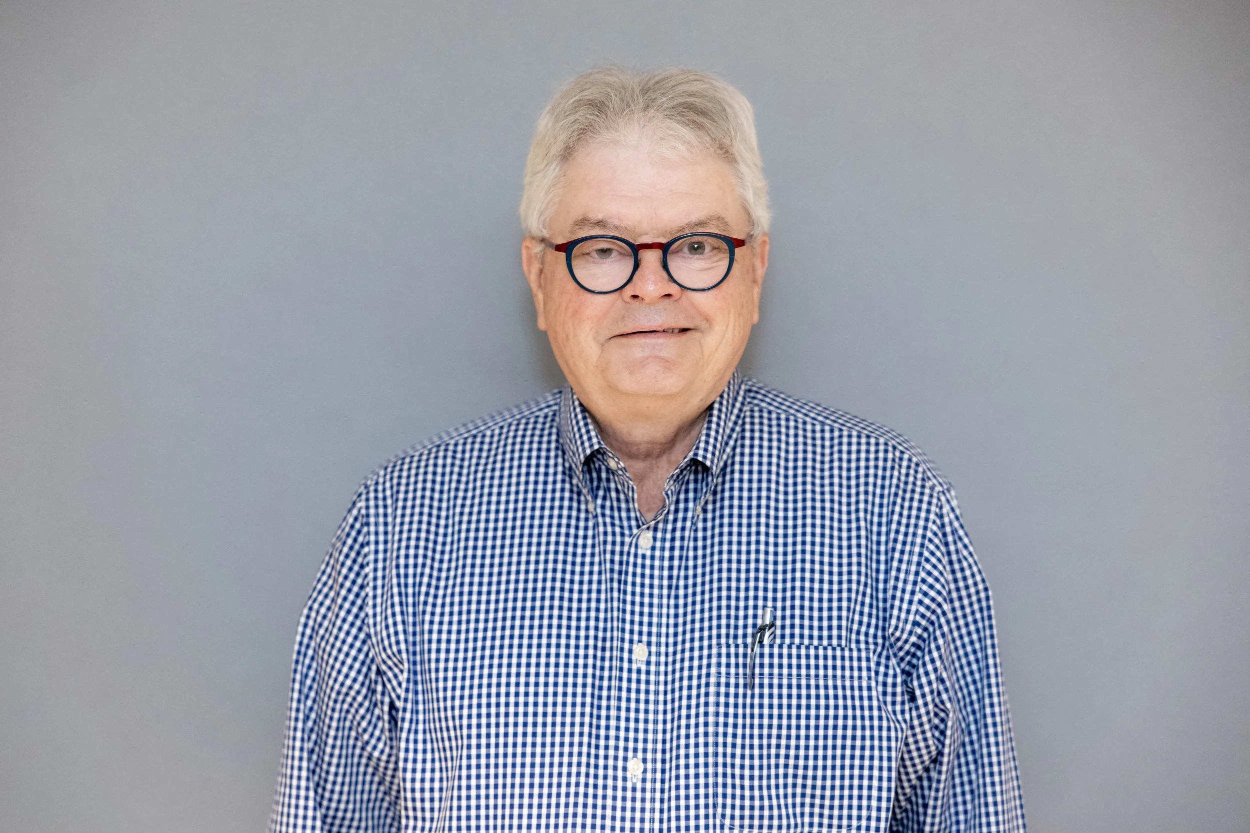 A middle-aged man with gray hair, wearing patterned glasses and a checkered button-down shirt, standing against a plain light gray background.