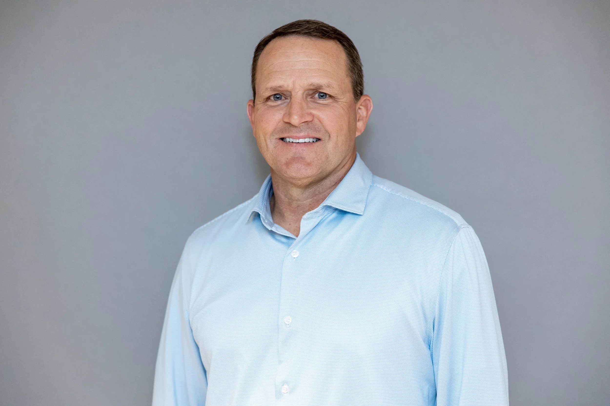 A middle-aged man with short brown hair, blue eyes, and wearing a light blue dress shirt, standing against a plain gray background, smiling at the camera.