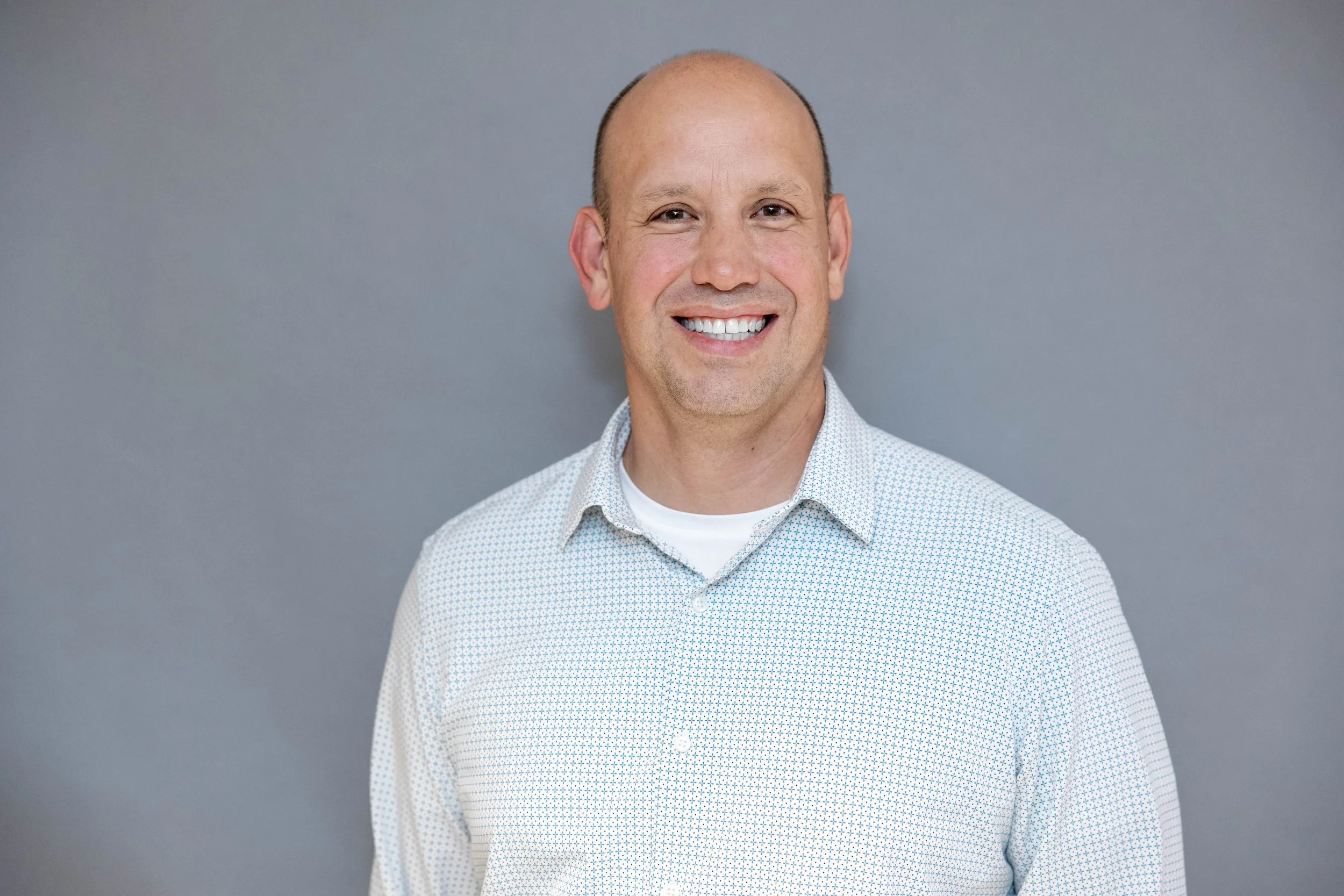 A smiling man with a shaved head, wearing a white, patterned button-up shirt, standing against a plain gray background.