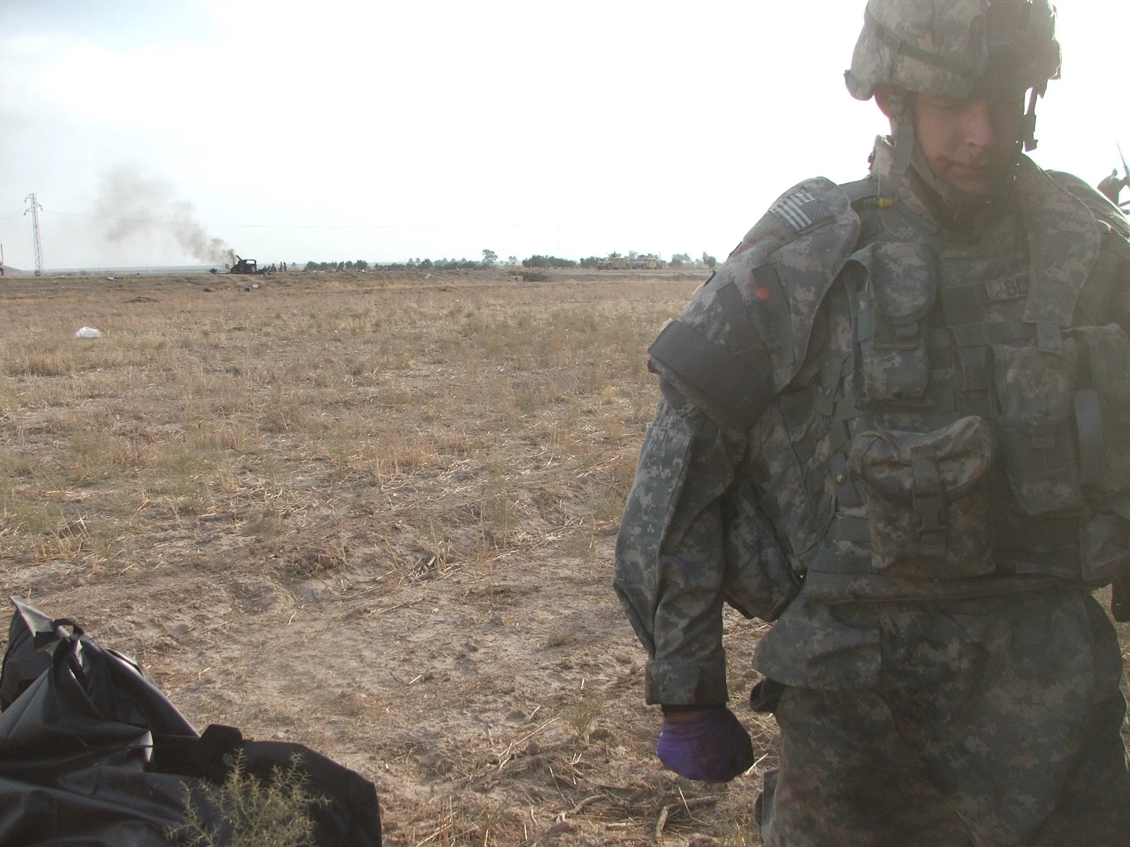 A soldier in camouflage military uniform and helmet standing in a dry, open field with smoke and a burning vehicle in the distance.